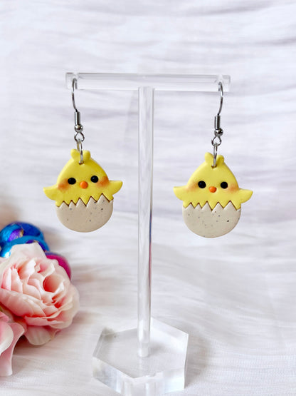 The Egg Baby Chick Polymer Clay Earrings by silverstar.nz feature a unique design of yellow chicks with small wings, orange beaks, and black eyes hatching from eggs on a clear stand. Pink roses and colorful fabric are in the background.