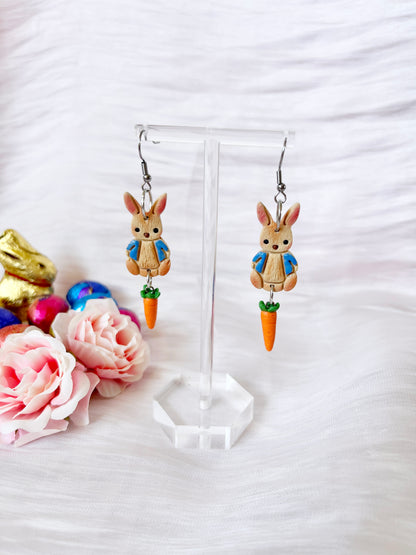 The Peter Rabbit Polymer Clay Earrings by silverstar.nz feature rabbits in blue jackets with orange carrots, displayed on a clear stand. Handmade from polymer clay with stainless steel hooks, they are whimsically set amidst pink roses and shiny egg-shaped decorations on a white textured surface.