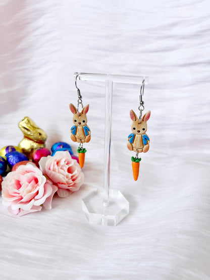 The Peter Rabbit Polymer Clay Earrings by silverstar.nz feature bunny figurines in blue jackets holding carrots, crafted from handmade polymer clay and suspended on stainless steel hooks. Displayed on a clear stand, they are surrounded by pastel Easter eggs, a gold bunny, and pink roses against a white textured background.