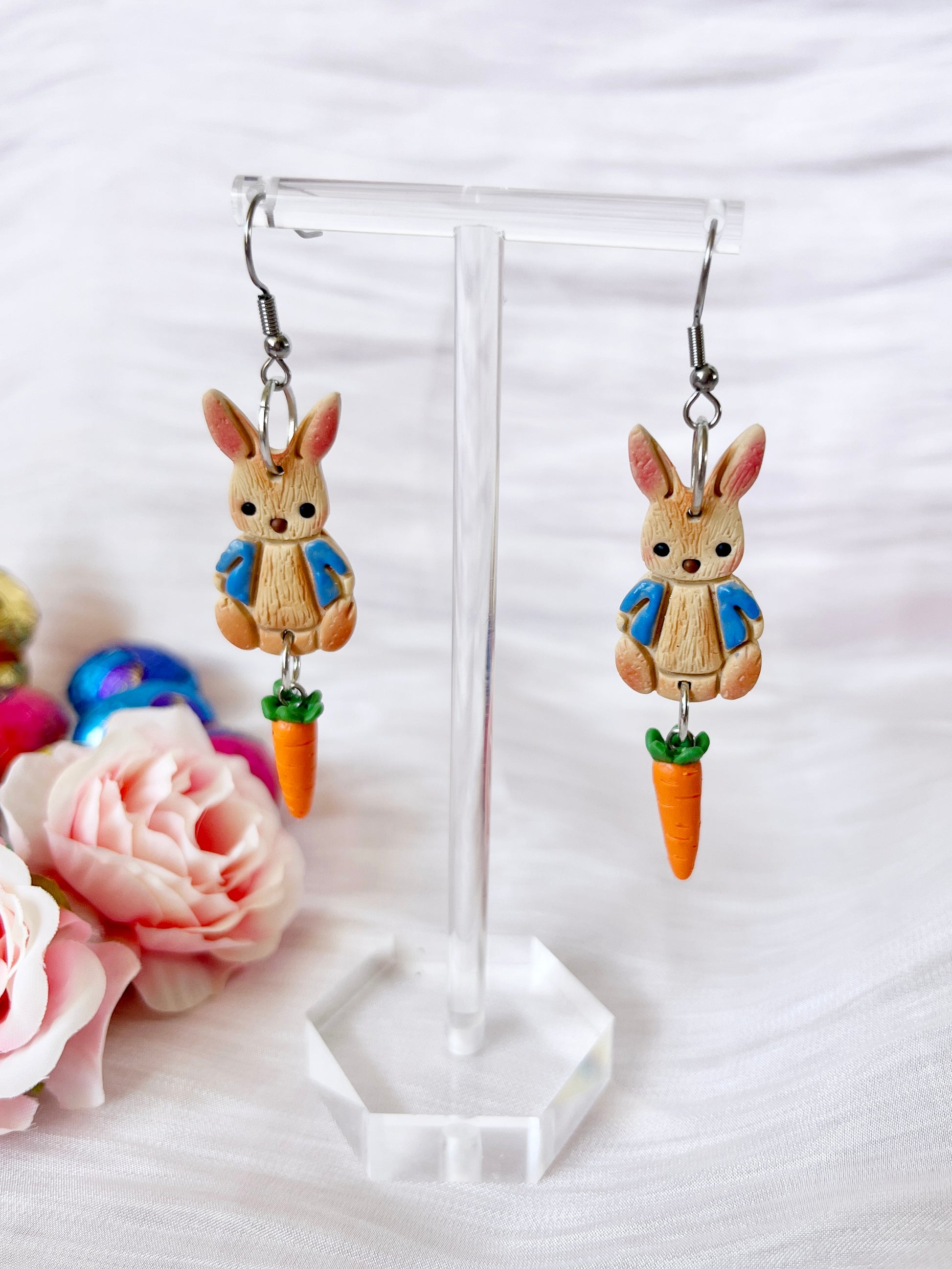 The Peter Rabbit Polymer Clay Earrings by silverstar.nz showcase bunny charms in blue jackets, each holding a carrot. Handmade from polymer clay, they are displayed with pink roses and colorful eggs. Stainless steel hooks complete the look.