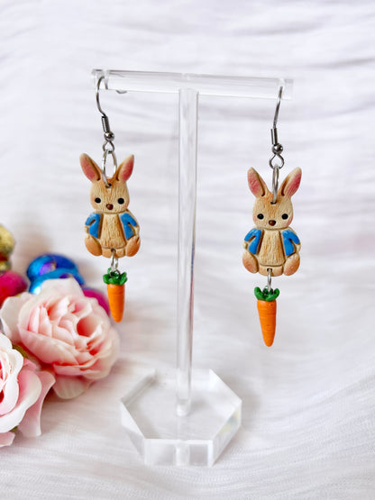 The Peter Rabbit Polymer Clay Earrings by silverstar.nz showcase bunny charms in blue jackets, each holding a carrot. Handmade from polymer clay, they are displayed with pink roses and colorful eggs. Stainless steel hooks complete the look.