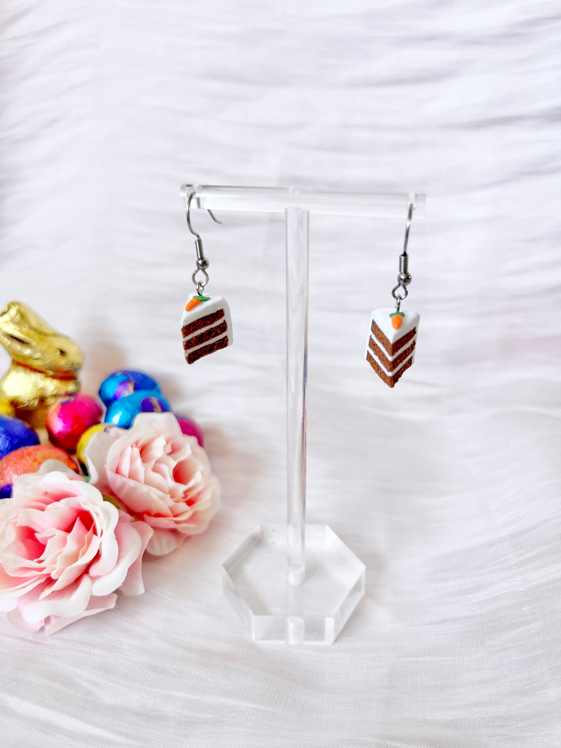 The Carrot Cake Polymer Clay Earrings by silverstar.nz dangle elegantly on a clear stand, resembling cake slices with white frosting. In the background, pink roses and colorful foil-wrapped chocolate eggs near a gold bunny enhance the charm of the white textured backdrop.