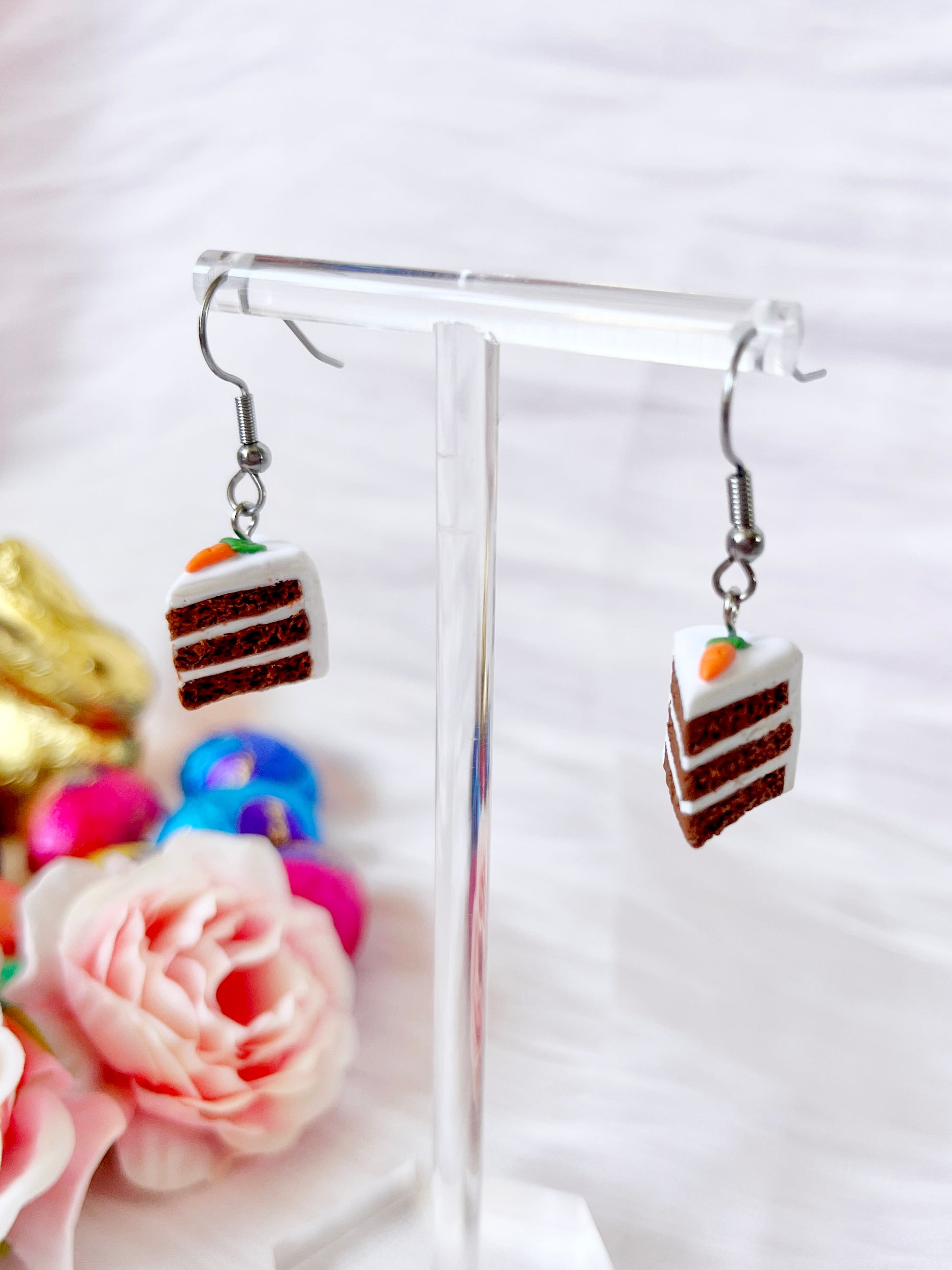 The Carrot Cake Polymer Clay Earrings by silverstar.nz are a funky, handmade design featuring chocolate cake slices with white frosting and carrot details, dangling elegantly on a clear stand against a rose-decorated backdrop.