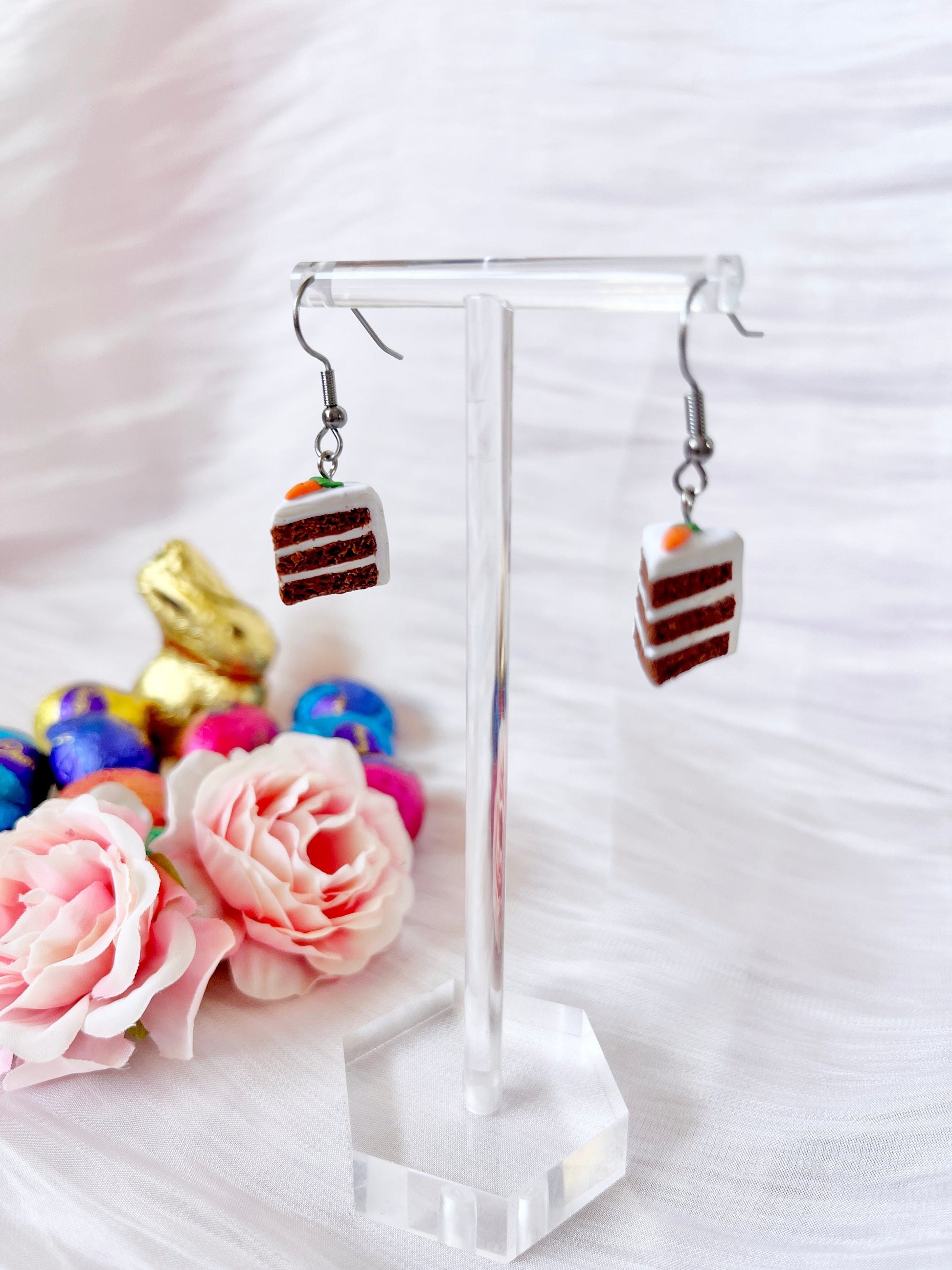 Carrot Cake Polymer Clay Earrings by silverstar.nz hang on a transparent stand amid pastel pink flowers and colorful chocolates, featuring a gold bunny, enhancing this whimsical display on the white textured surface.