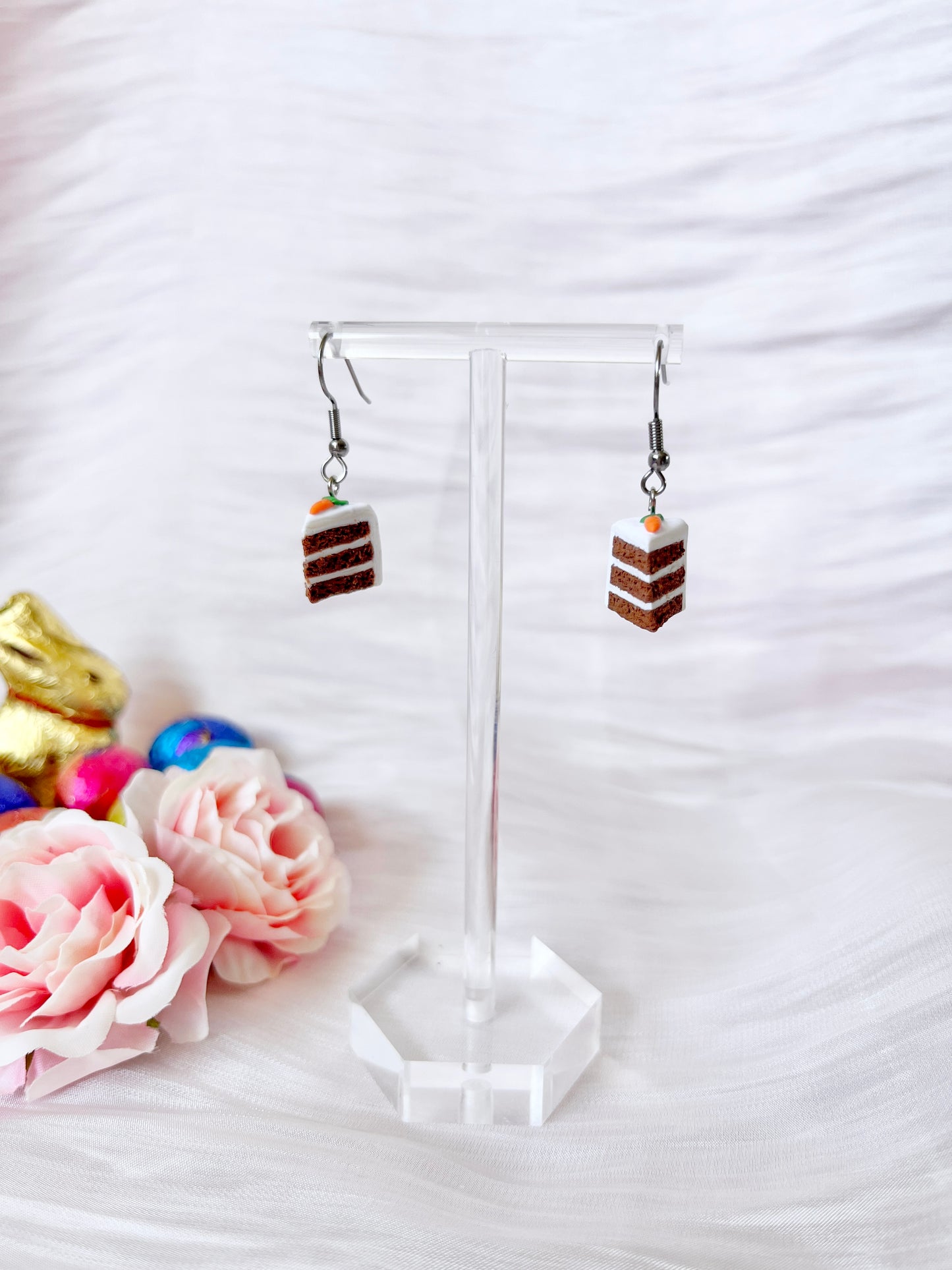The Carrot Cake Polymer Clay Earrings by silverstar.nz dangle gracefully on a clear stand, adorned with pink flowers and colorful Easter eggs, set against a soft, white textured fabric.
