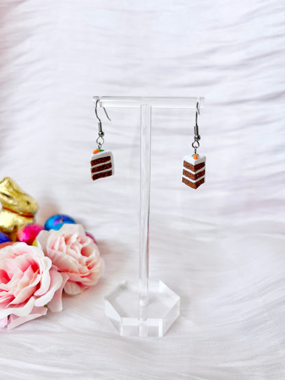 The Carrot Cake Polymer Clay Earrings by silverstar.nz dangle gracefully on a clear stand, adorned with pink flowers and colorful Easter eggs, set against a soft, white textured fabric.