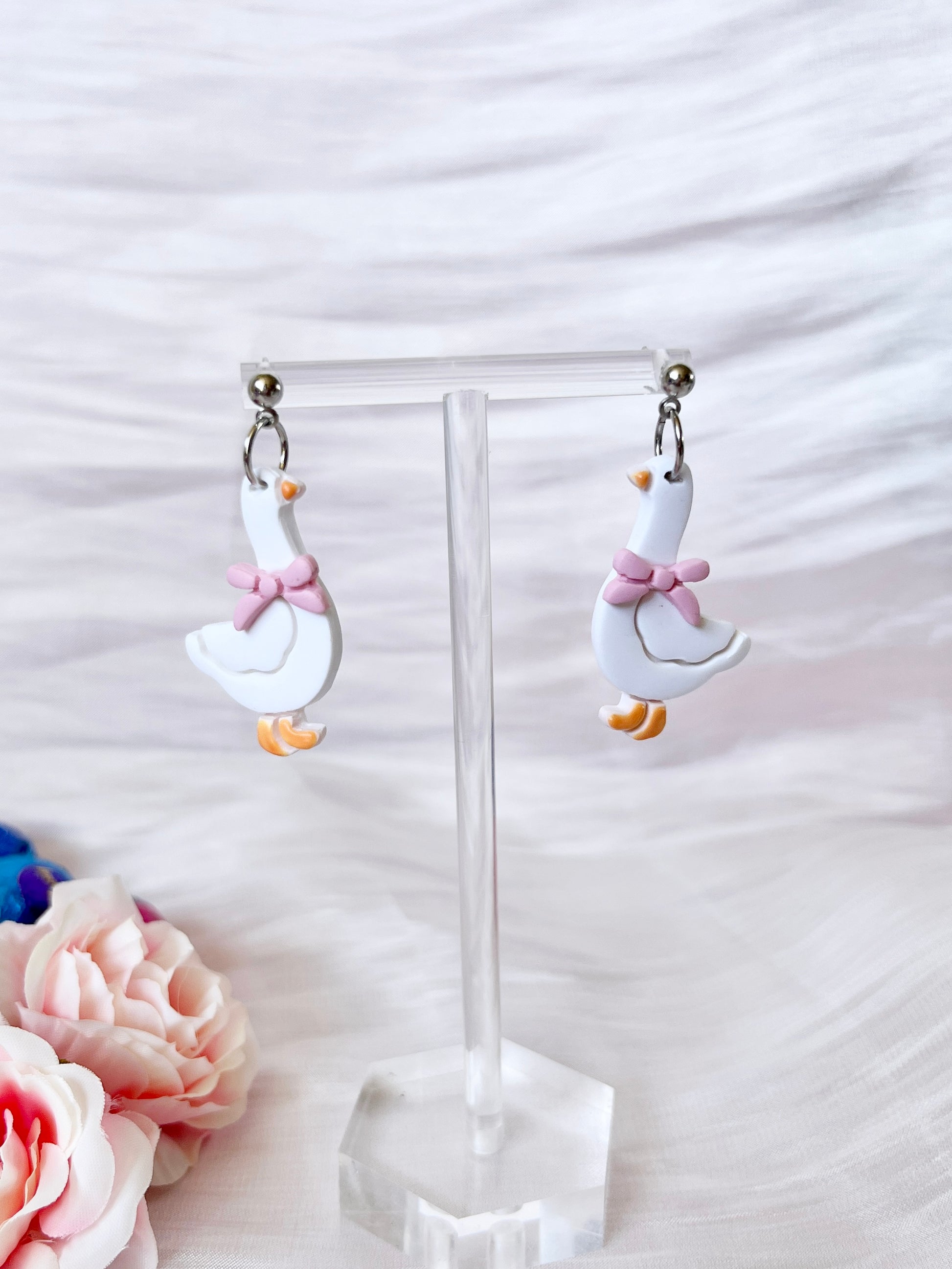 Goose with Bow Polymer Clay Earrings by silverstar.nz—handmade white goose-shaped earrings featuring orange beaks, feet, and pink bows; displayed on a clear stand with soft white fabric and pink flowers for a charming, unique look.