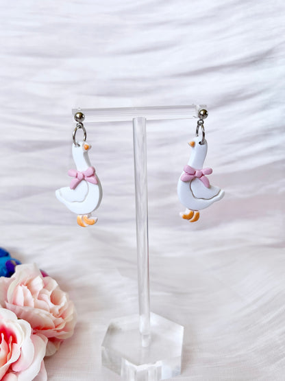 Goose with Bow Polymer Clay Earrings by silverstar.nz—handmade white goose-shaped earrings featuring orange beaks, feet, and pink bows; displayed on a clear stand with soft white fabric and pink flowers for a charming, unique look.