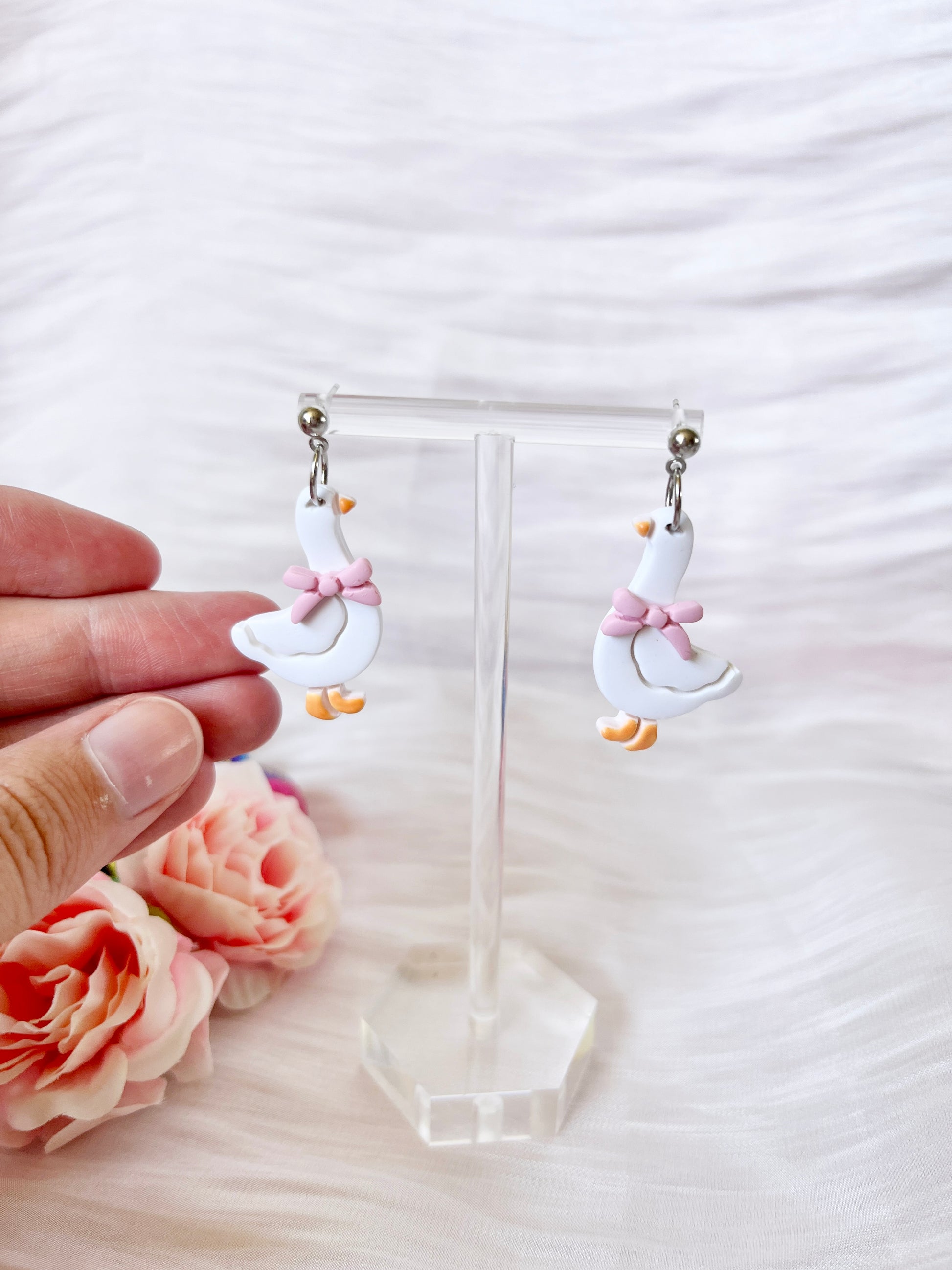Goose with Bow Polymer Clay Earrings by silverstar.nz are showcased on a clear stand, with a hand adjusting one. Pink flowers decorate the soft, light fabric background.