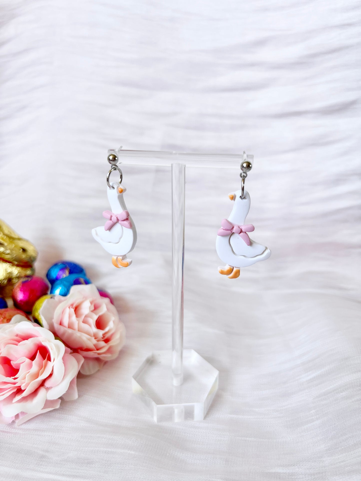 Goose with Bow Polymer Clay Earrings by silverstar.nz—white goose-shaped, handmade earrings with pink bows—are shown on a clear stand amid pink roses and colorful eggs on soft white fabric.