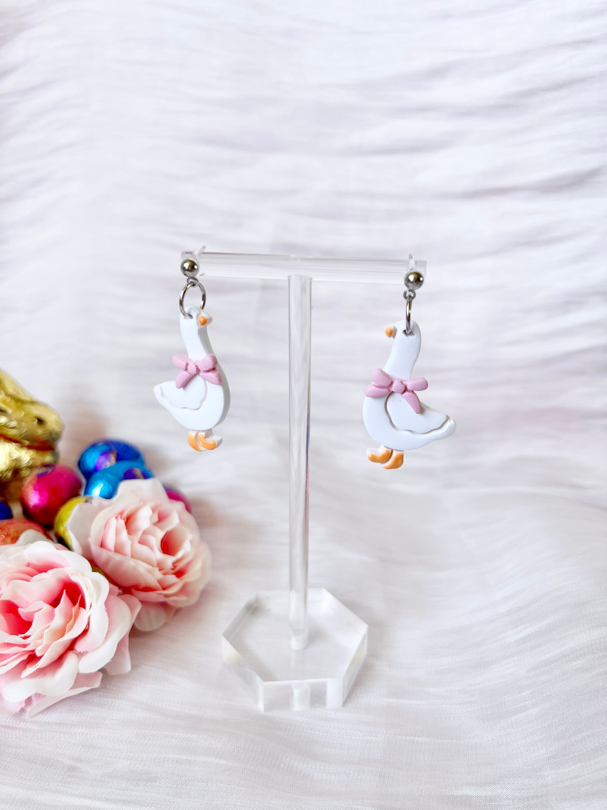 Goose with Bow Polymer Clay Earrings by silverstar.nz—white goose-shaped, handmade earrings with pink bows—are shown on a clear stand amid pink roses and colorful eggs on soft white fabric.