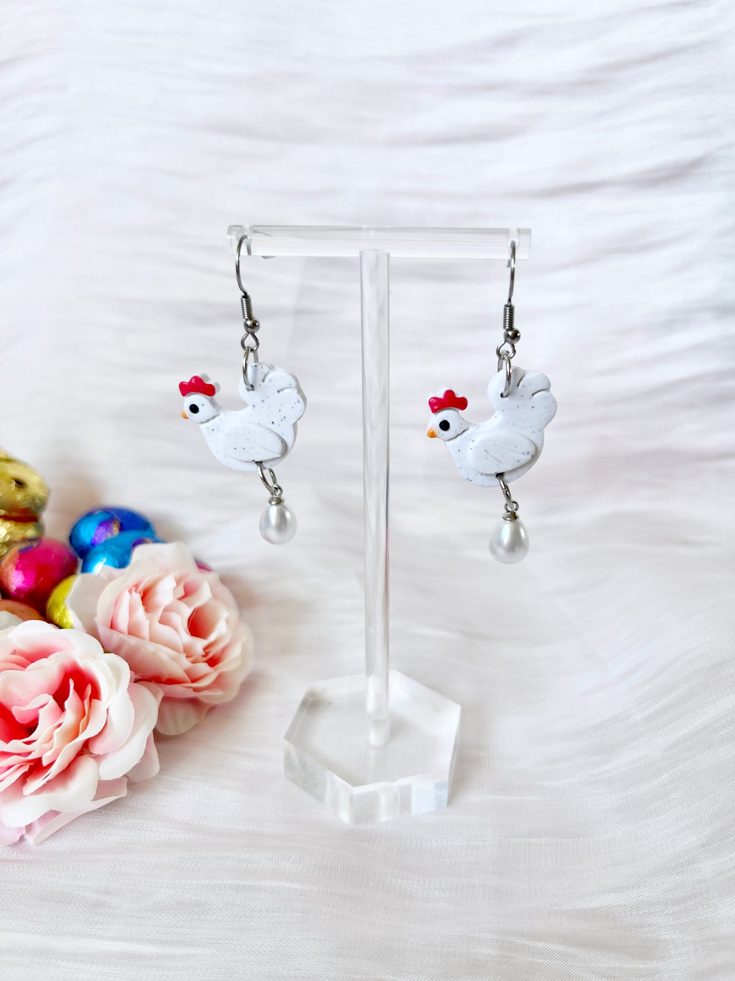 The Chicken with Egg Polymer Clay Earrings by silverstar.nz feature white chicken shapes with red combs and pearl drops, displayed on stainless steel hooks against a textured white background with pink roses and foil-wrapped chocolates.