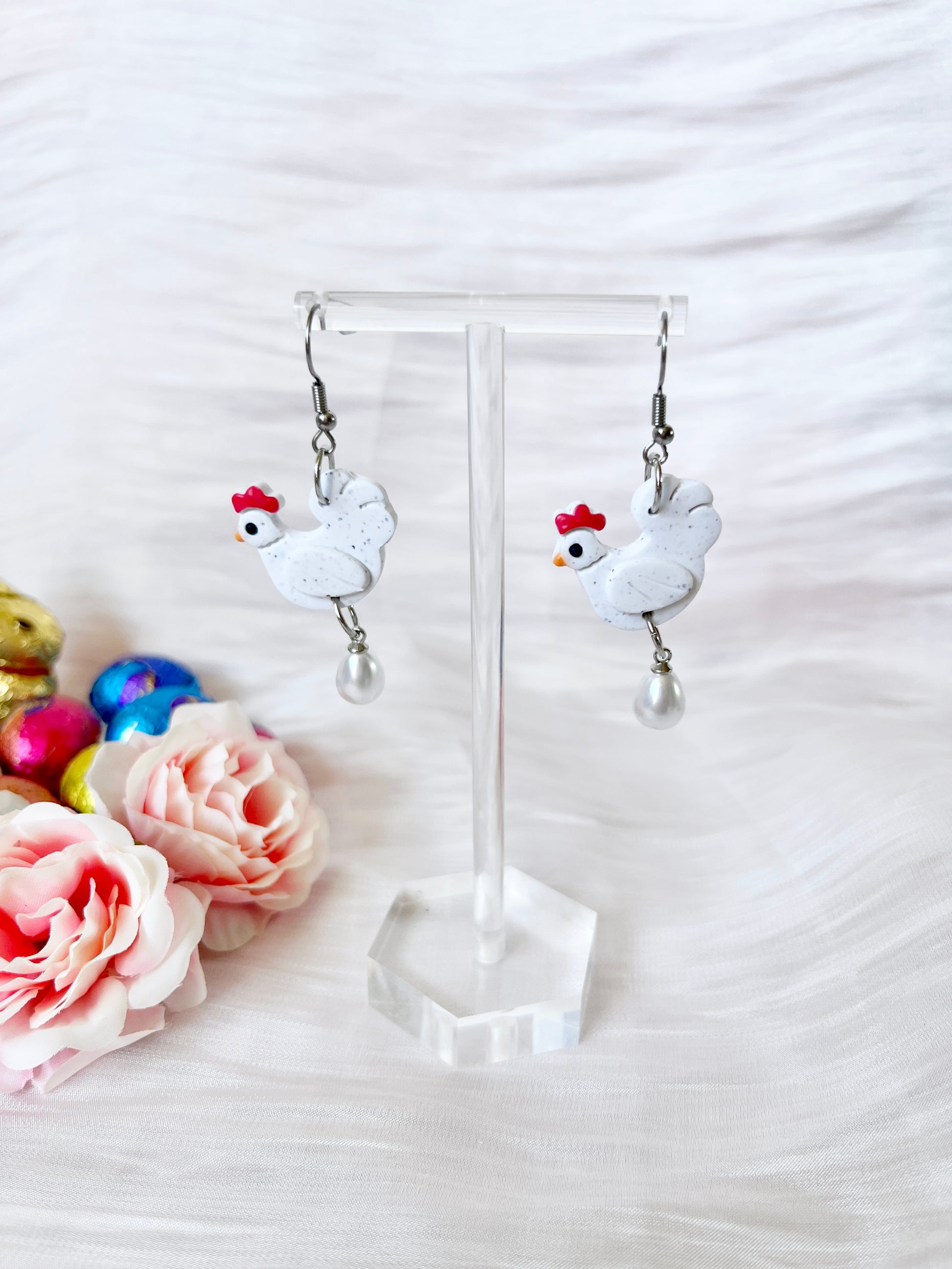 The Chicken with Egg Polymer Clay Earrings by silverstar.nz feature white chicken shapes with red combs and pearl drops, displayed on stainless steel hooks against a textured white background with pink roses and foil-wrapped chocolates.