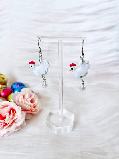 The Chicken with Egg Polymer Clay Earrings by silverstar.nz feature white chicken shapes with red combs and pearl drops, displayed on stainless steel hooks against a textured white background with pink roses and foil-wrapped chocolates.