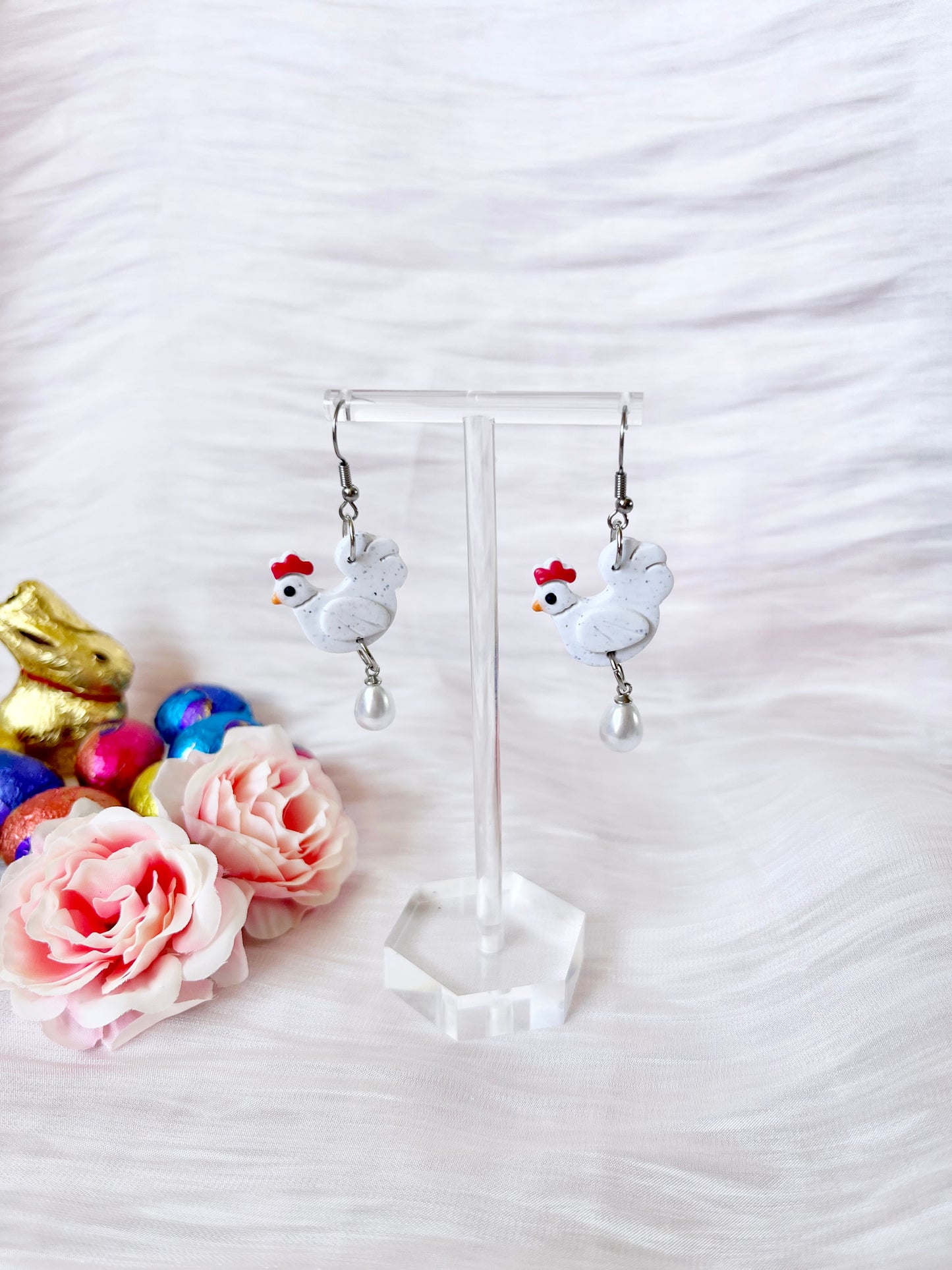 A pair of silverstar.nz Chicken with Egg Polymer Clay Earrings with stainless steel hooks and pearl drops is displayed on a clear stand, surrounded by pink faux flowers, chocolate bunnies, and colorful Easter eggs against a white textured background.