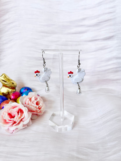 A pair of silverstar.nz Chicken with Egg Polymer Clay Earrings with stainless steel hooks and pearl drops is displayed on a clear stand, surrounded by pink faux flowers, chocolate bunnies, and colorful Easter eggs against a white textured background.