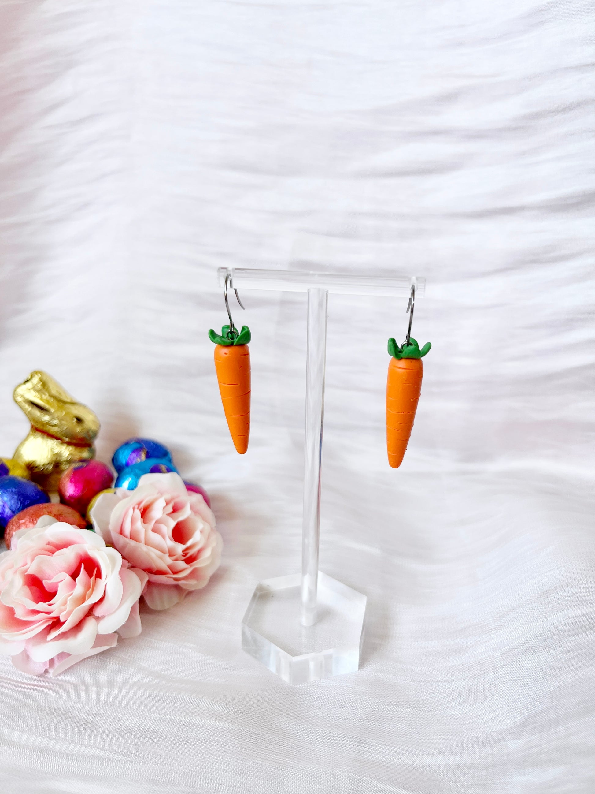 Two carrot-shaped earrings with green tops by silverstar.nz hang on a transparent stand. Pink roses, a golden rabbit figure, and colorful eggs decorate the white surface in the background. These handmade Carrot Polymer Clay Earrings feature a unique design that catches every eye.