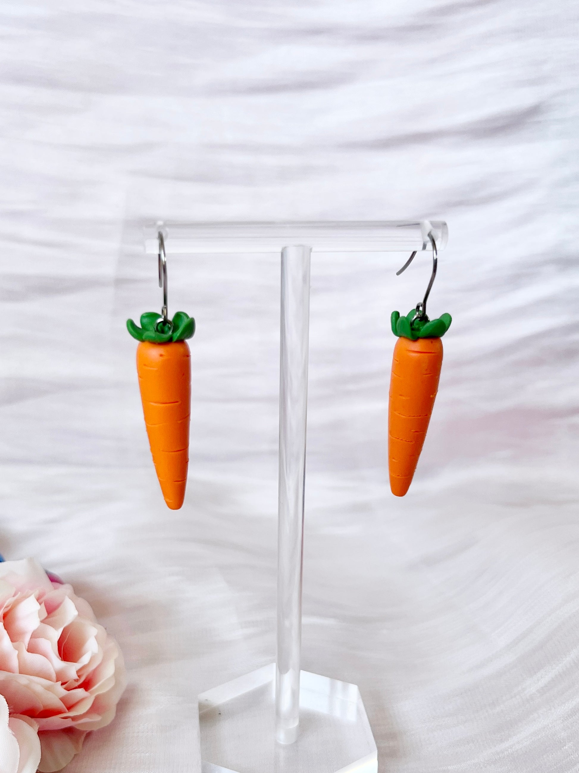 The Carrot Polymer Clay Earrings by silverstar.nz are showcased on a clear stand, set against a soft, light pink and white striped background. A pale pink rose is partially visible in the bottom left corner, adding to the handcrafted charm of these unique earrings designed as carrots with green tops.