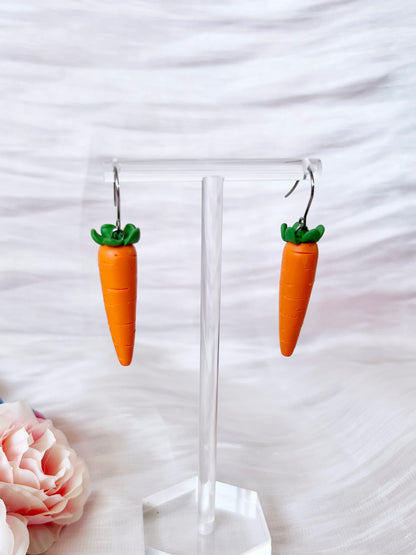 The Carrot Polymer Clay Earrings by silverstar.nz are showcased on a clear stand, set against a soft, light pink and white striped background. A pale pink rose is partially visible in the bottom left corner, adding to the handcrafted charm of these unique earrings designed as carrots with green tops.
