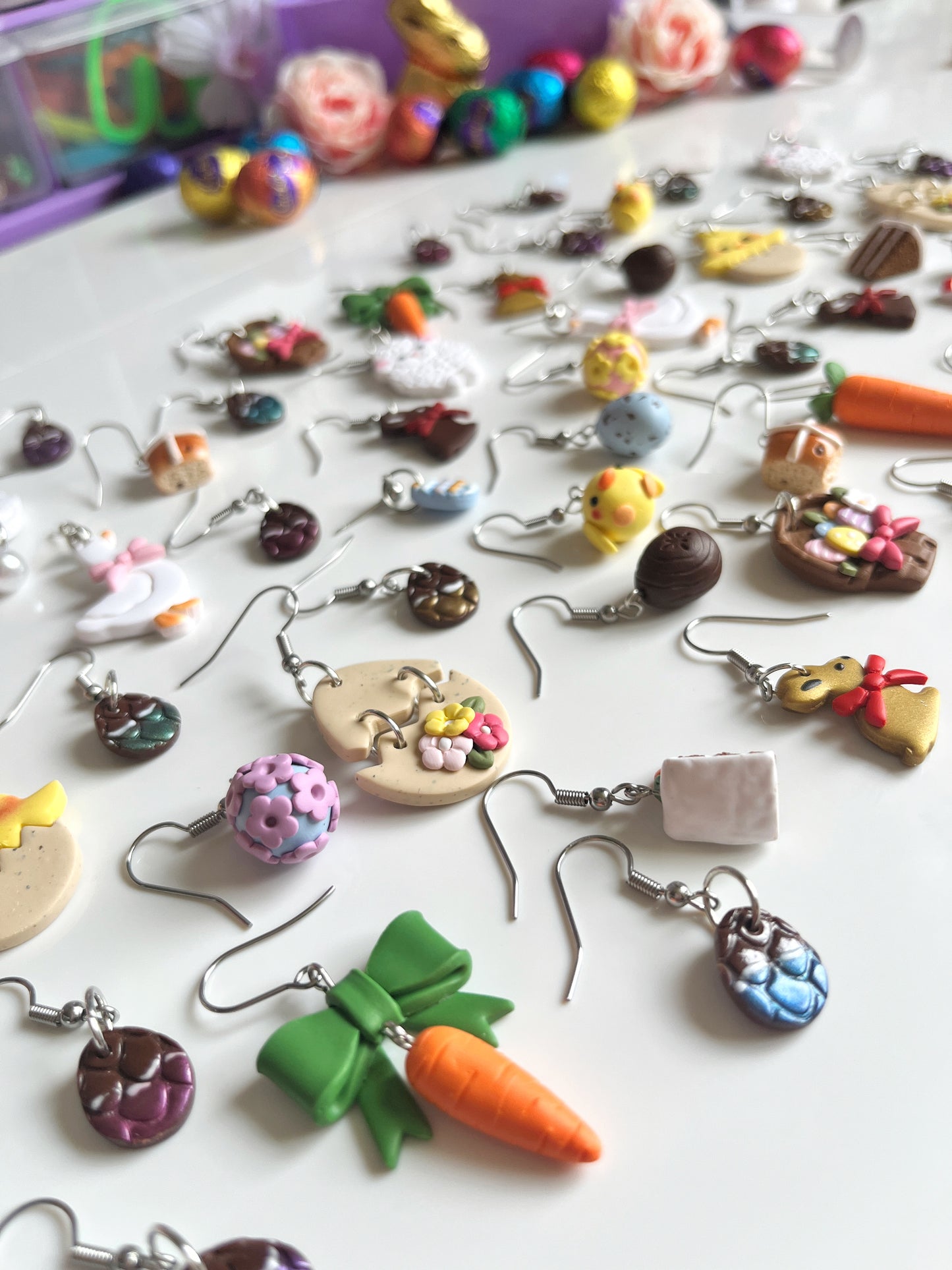 A colorful set of silverstar.nzs handmade polymer clay earrings is displayed on a white surface. The unique designs include Carrots with a Bow, cookies, and chicks, featuring a playful, crafty theme. Blurred colorful objects are visible in the background.