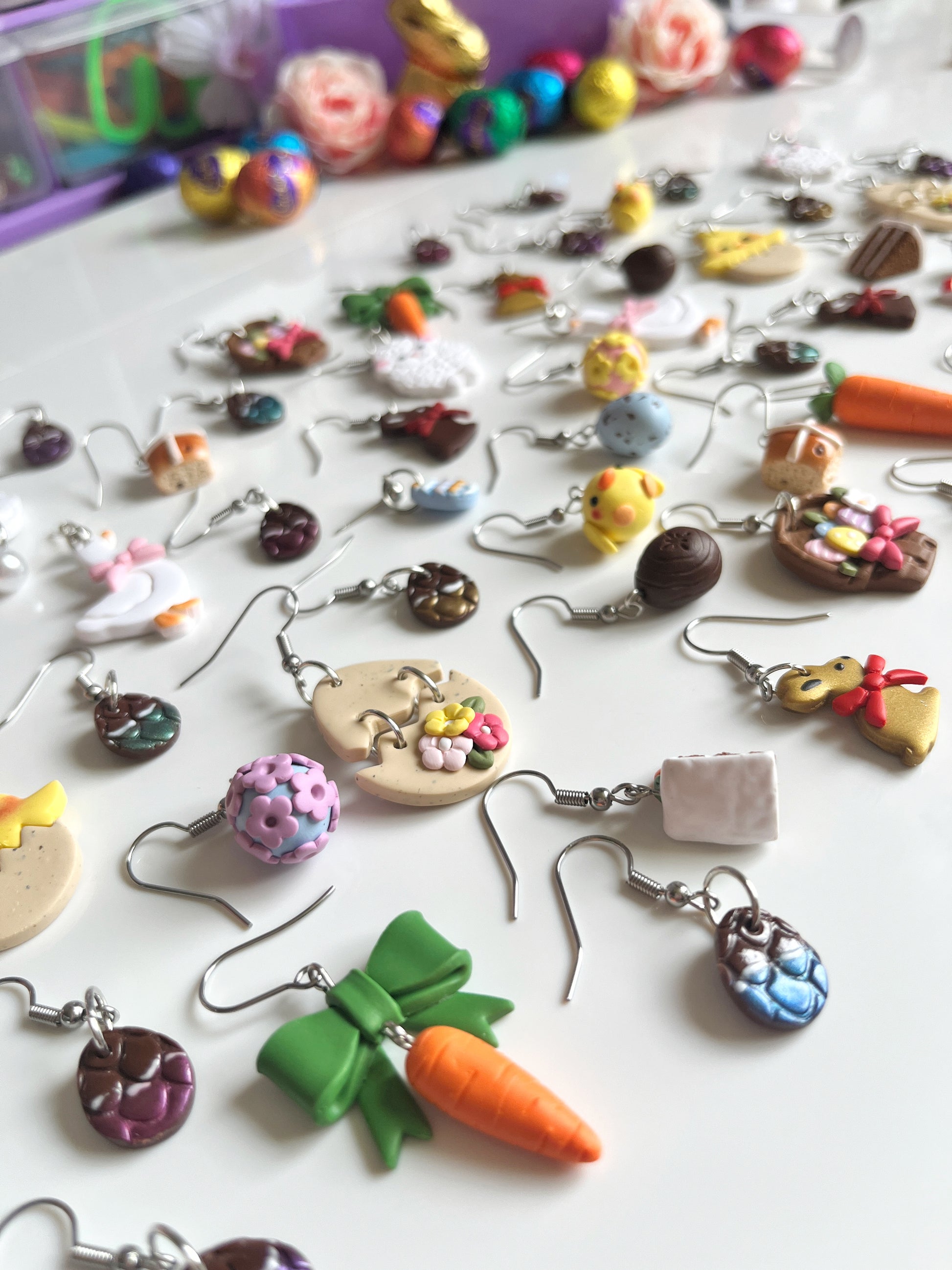 A colorful set of silverstar.nzs handmade polymer clay earrings is displayed on a white surface. The unique designs include Carrots with a Bow, cookies, and chicks, featuring a playful, crafty theme. Blurred colorful objects are visible in the background.