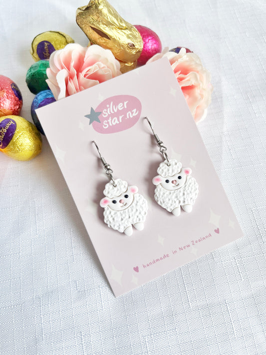 The Fluffy Sheep Polymer Clay Earrings by silverstar.nz are showcased on a pink card. Displayed on a textured white surface, these handmade earrings feature vibrant foil-wrapped eggs and pastel flowers in the background, emphasizing their unique design.