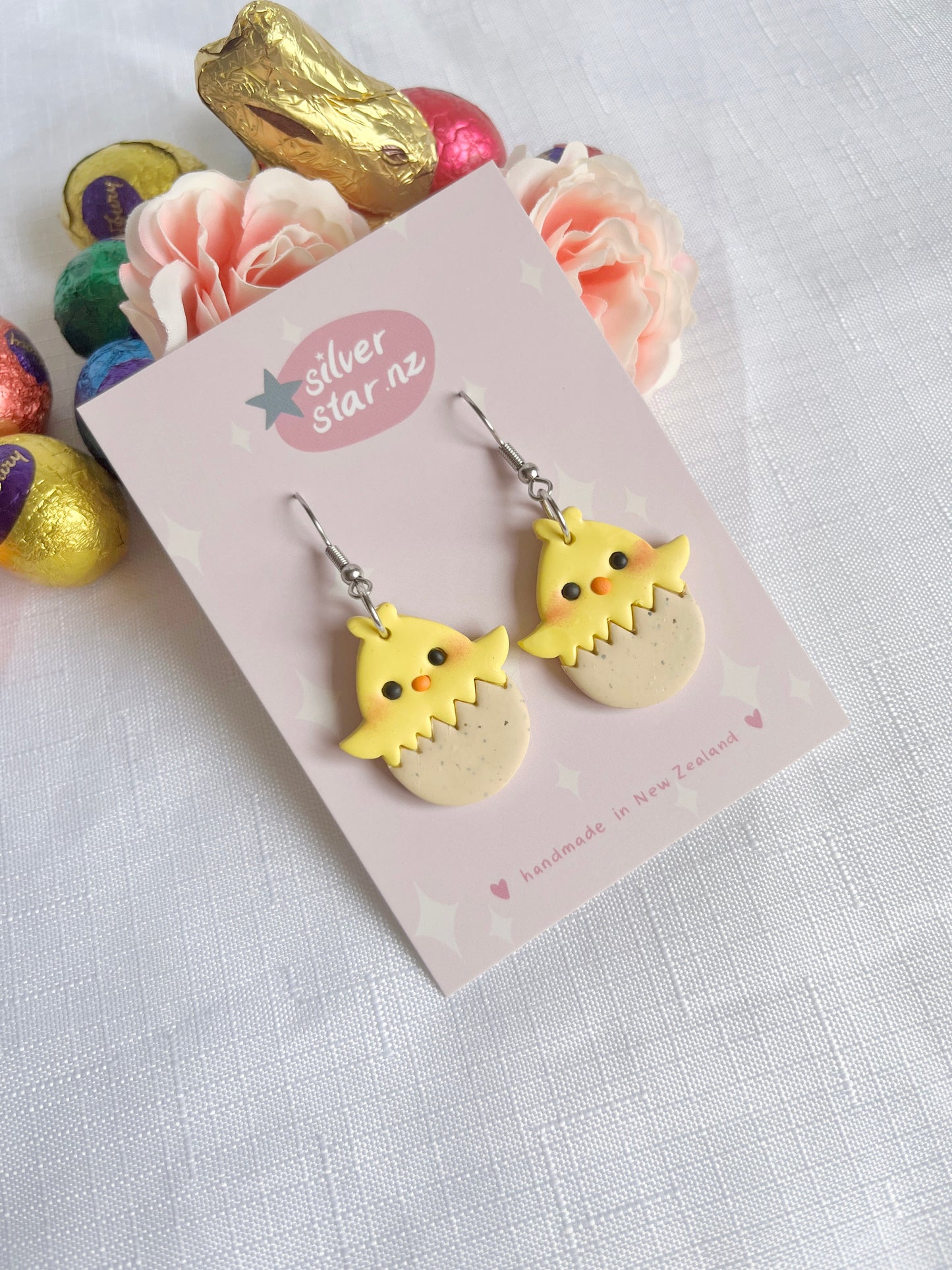 Egg Baby Chick Polymer Clay Earrings by silverstar.nz are uniquely designed with yellow chicks hatching from eggs, displayed on a silverstar.nz card. Theyre surrounded by chocolate eggs and peach roses on a white textured cloth.
