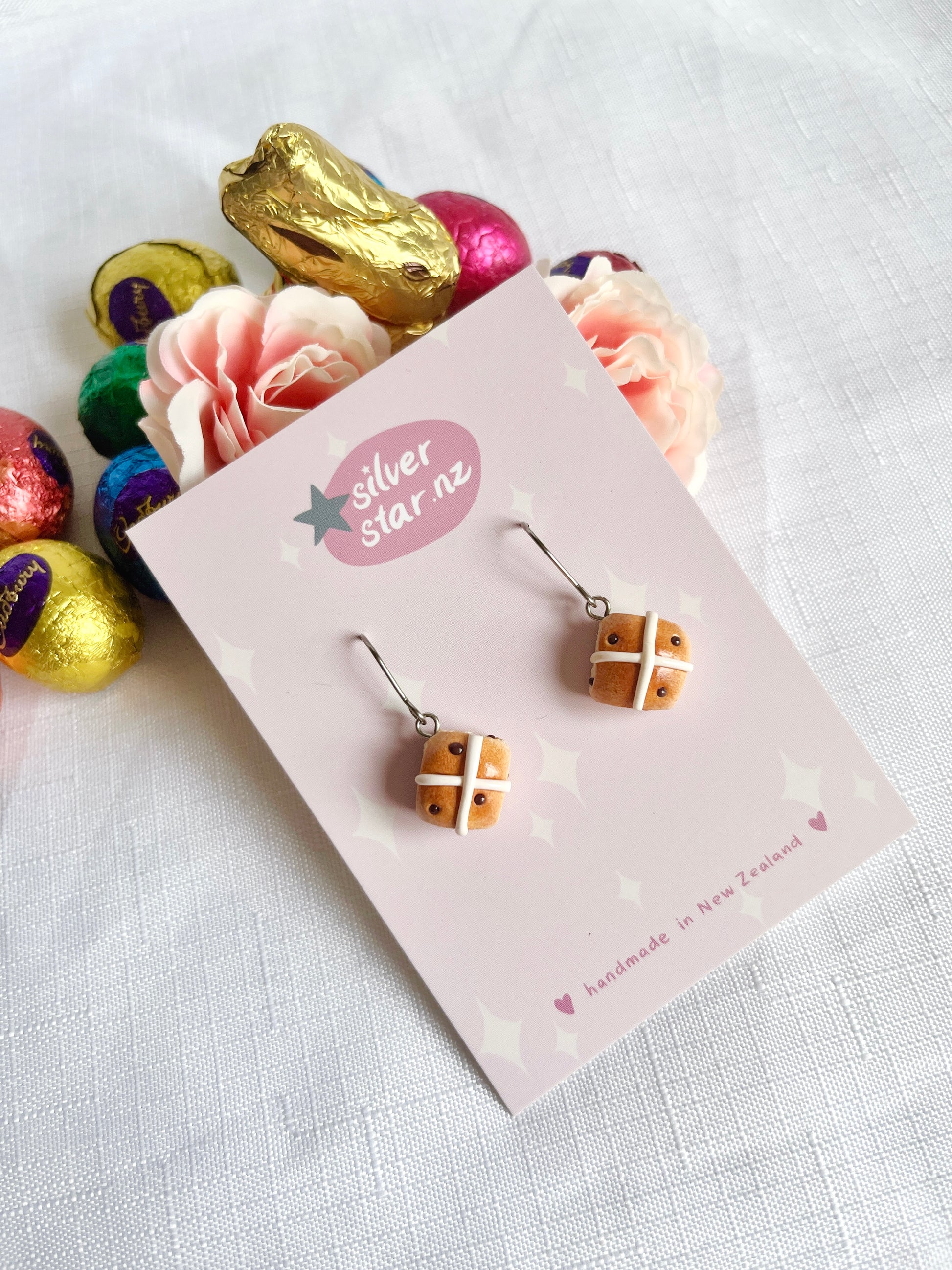 A set of handmade Hot Cross Bun Polymer Clay Earrings on a silverstar.nz card is complemented by a faux gold-wrapped bunny and vibrant chocolate eggs. Soft pink roses and a white cloth in the background enhance these unique design earrings with stainless steel hooks.