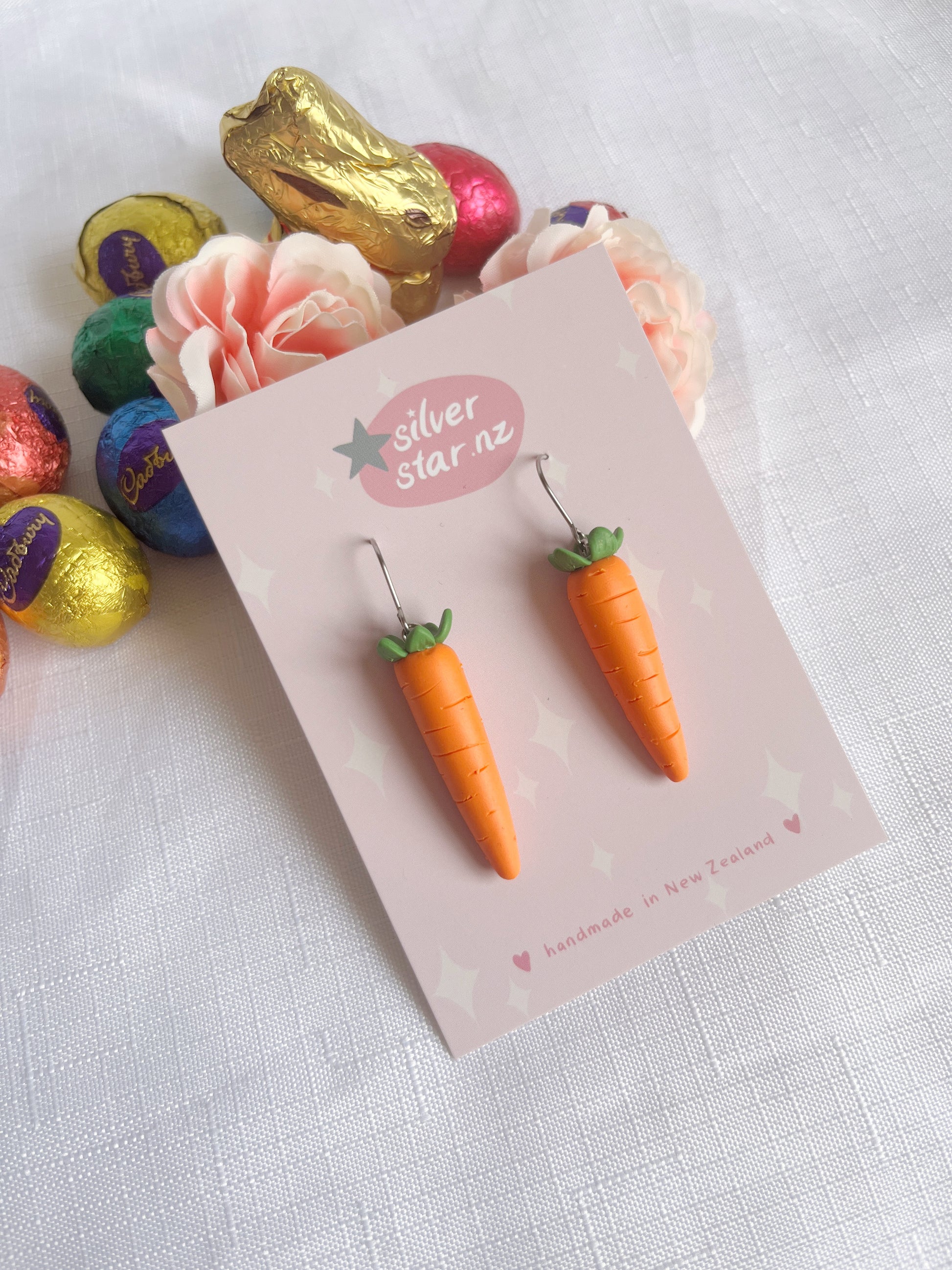 The Carrot Polymer Clay Earrings by silverstar.nz are artfully displayed on a card. Situated among vibrant foil-wrapped eggs and a golden lamb on a white textured surface, they lie beside a delicate light pink flower, highlighting their unique design.