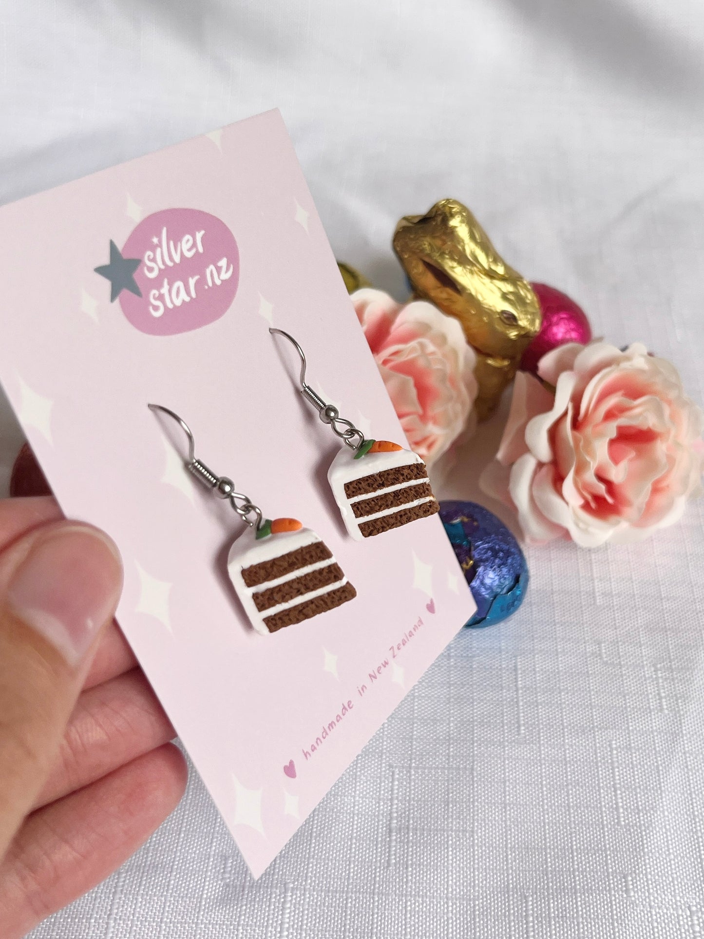 A hand holds Carrot Cake Polymer Clay Earrings on a silverstar.nz card with stainless steel hooks, amid pastel roses and foil-wrapped chocolate eggs on white fabric.