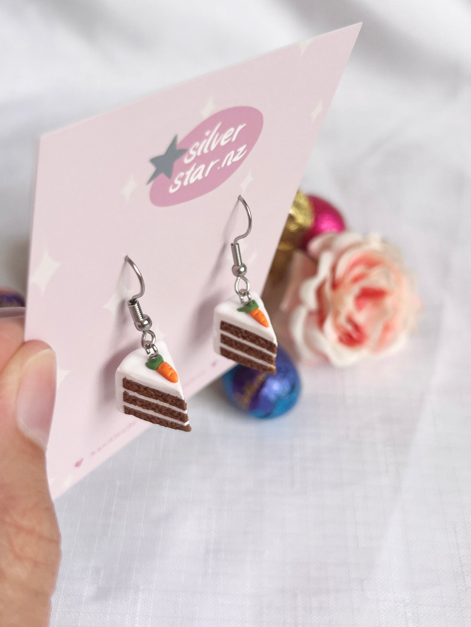 A hand displays a card featuring silverstar.nzs Carrot Cake Polymer Clay Earrings, artistically crafted to resemble carrot cake slices with white frosting and a tiny carrot. A blurred pink rose and colorful decorative balls in the background enhance the funky charm.