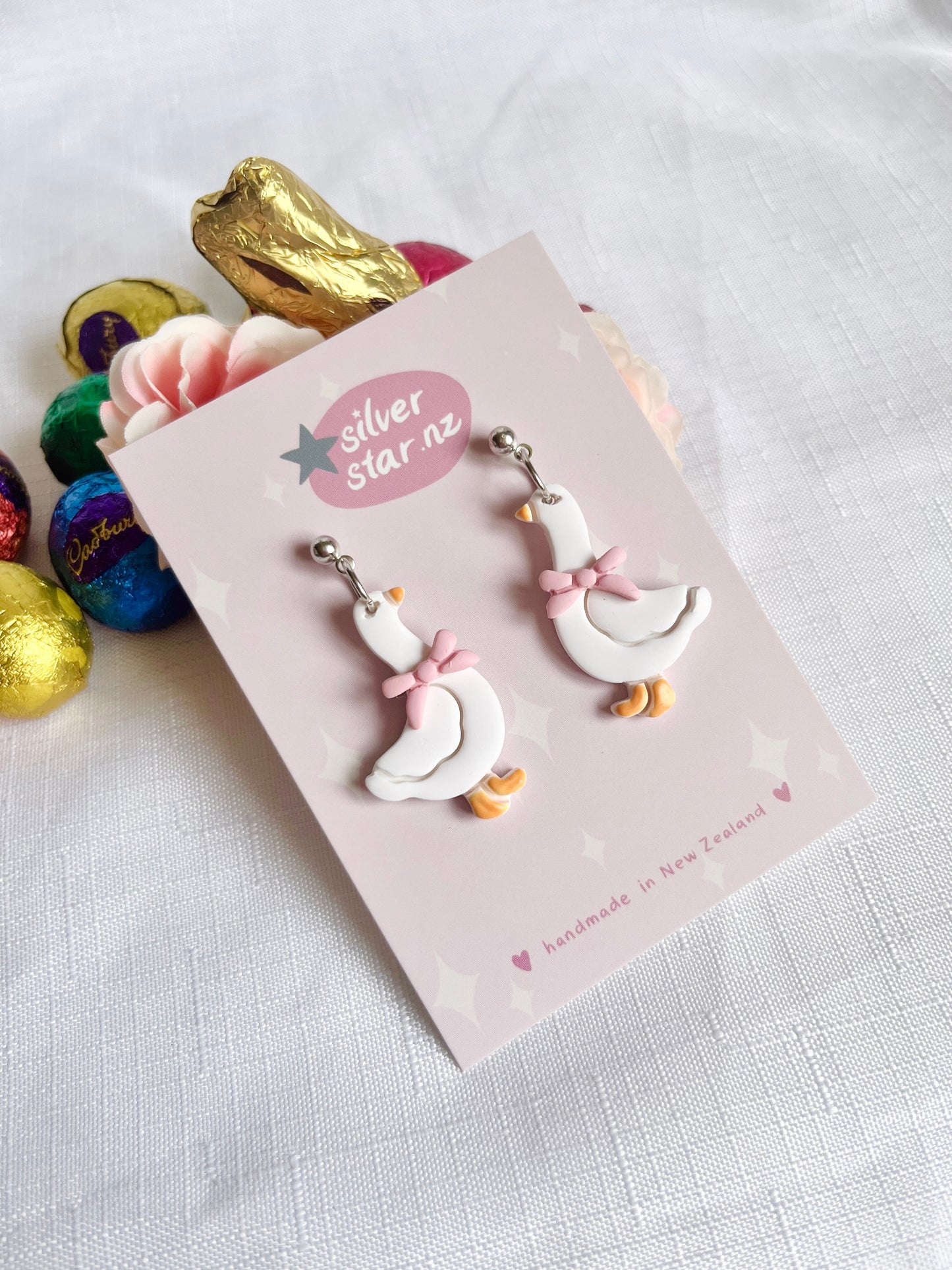 Goose with Bow Polymer Clay Earrings by silverstar.nz, handmade in New Zealand. Featuring a unique white goose design with pink bows, displayed on a branded card. Background includes colorful foil-wrapped chocolate eggs and a pink rose on white fabric.