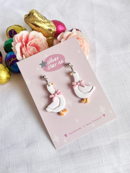 Goose with Bow Polymer Clay Earrings by silverstar.nz, featuring a unique design, are handmade and displayed on a silver star NZ card near pink flowers, a gold-wrapped chocolate bunny, and colorful chocolate eggs on white fabric.