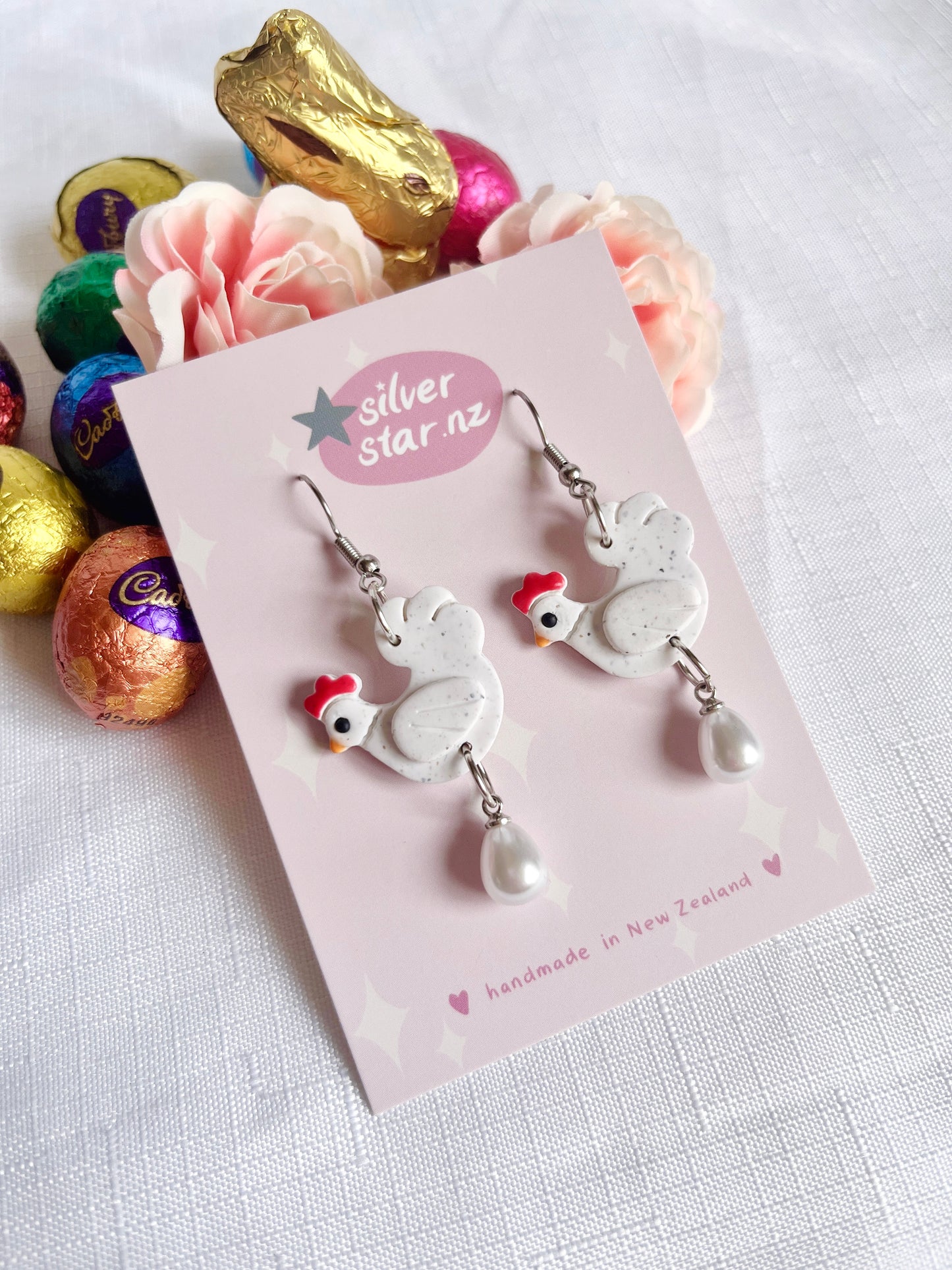 Chicken with Egg Polymer Clay Earrings by silverstar.nz: Handmade earrings shaped like chickens with red combs and dangling pearls, displayed on a pink card and surrounded by pastel flowers and colorful chocolate eggs.
