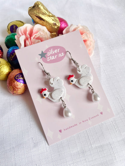 Chicken with Egg Polymer Clay Earrings by silverstar.nz: Handmade earrings shaped like chickens with red combs and dangling pearls, displayed on a pink card and surrounded by pastel flowers and colorful chocolate eggs.