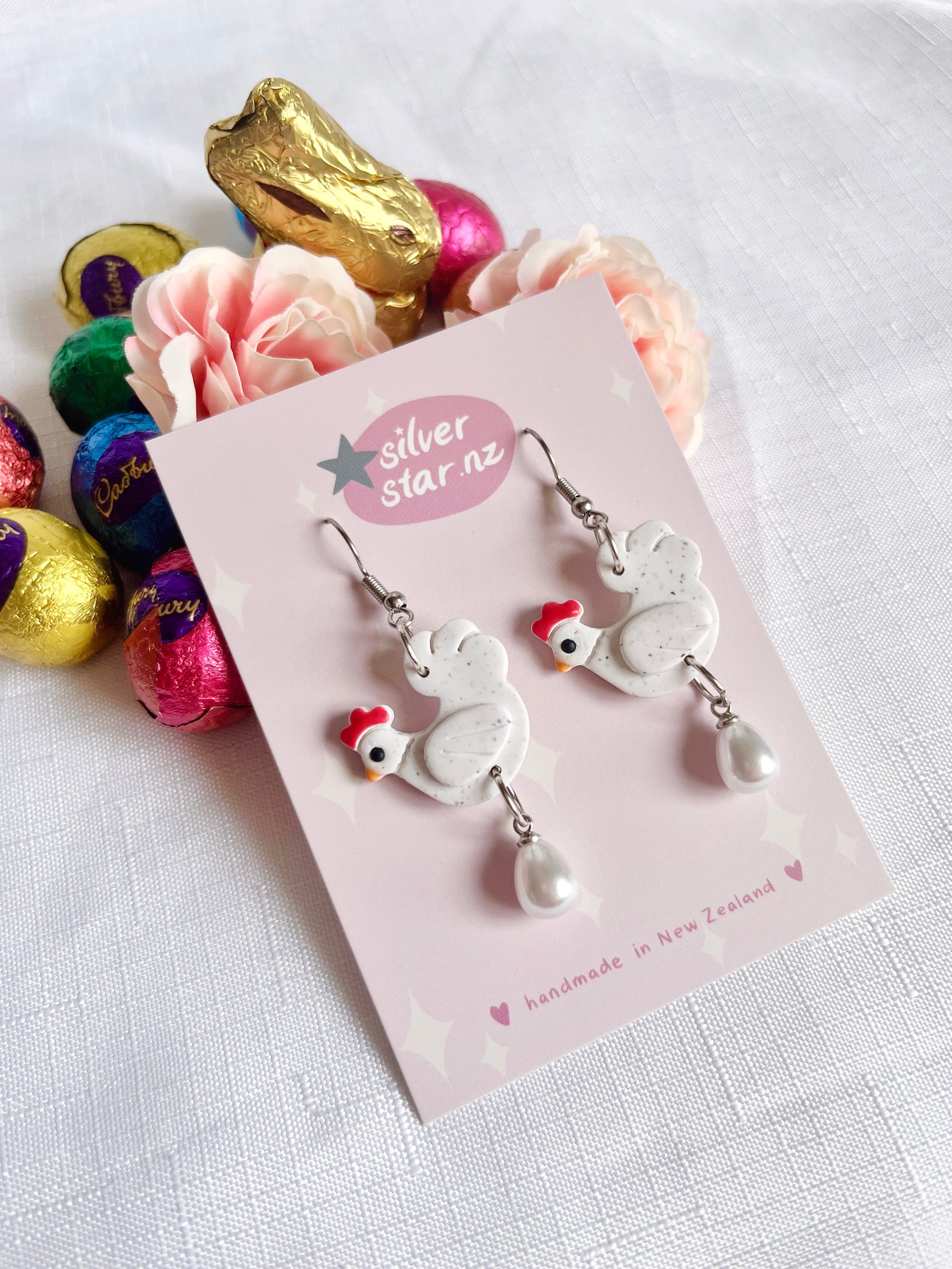 Chicken with Egg Polymer Clay Earrings by silverstar.nz—handmade chicken-shaped earrings with pearl drops and stainless steel hooks, shown on a pink card amid artificial flowers and colorful foil-wrapped chocolate eggs.