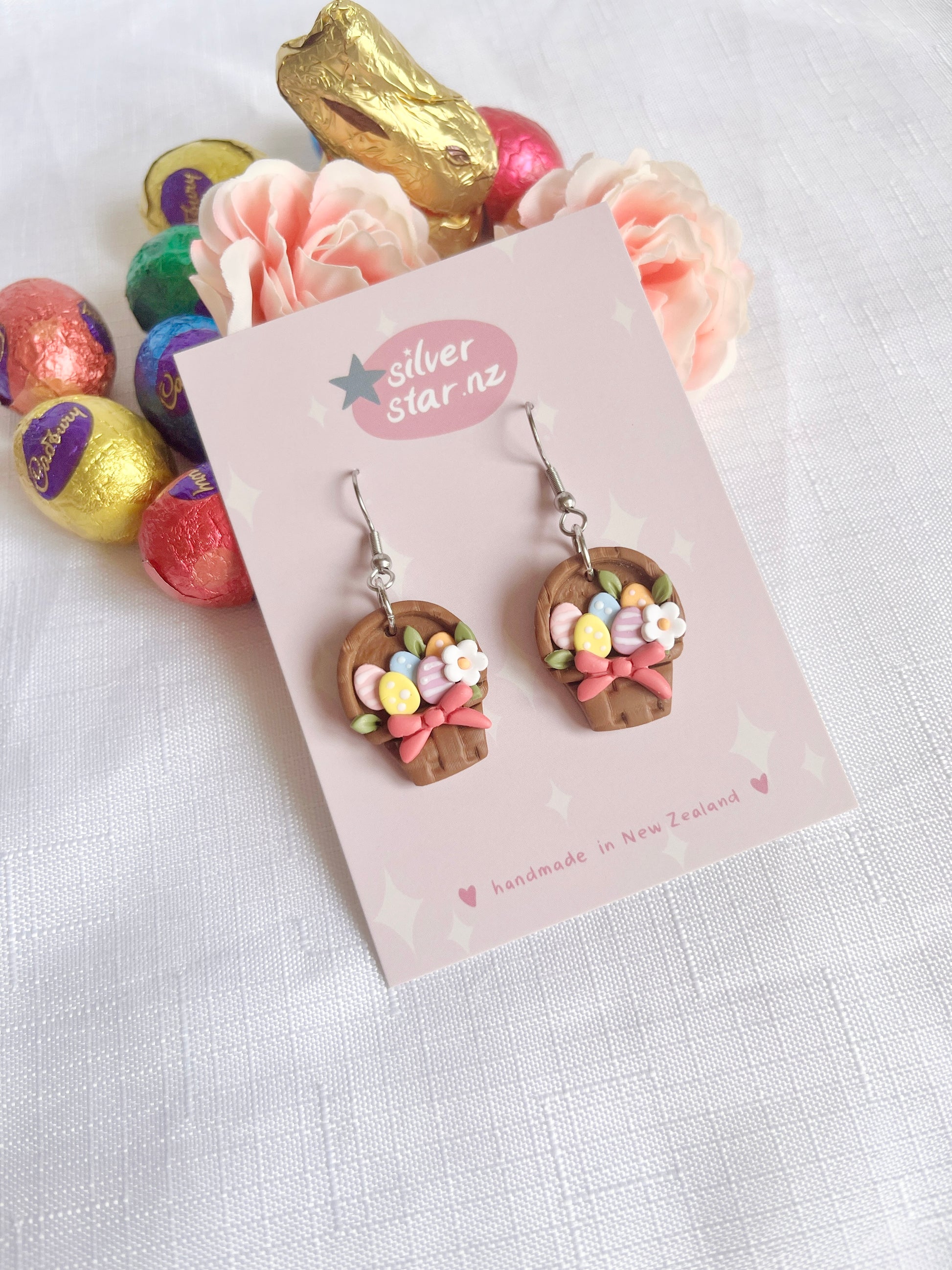 Egg Basket Polymer Clay Earrings from silverstar.nz, shaped like Easter baskets with colorful eggs and bows, are displayed on a card. Theyre surrounded by pink roses and foil-wrapped chocolate eggs on a white cloth.