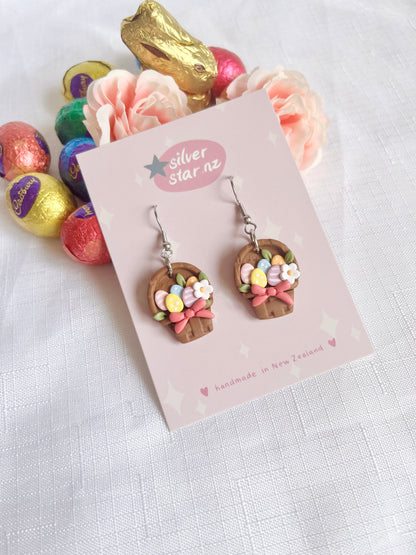 Egg Basket Polymer Clay Earrings from silverstar.nz, shaped like Easter baskets with colorful eggs and bows, are displayed on a card. Theyre surrounded by pink roses and foil-wrapped chocolate eggs on a white cloth.