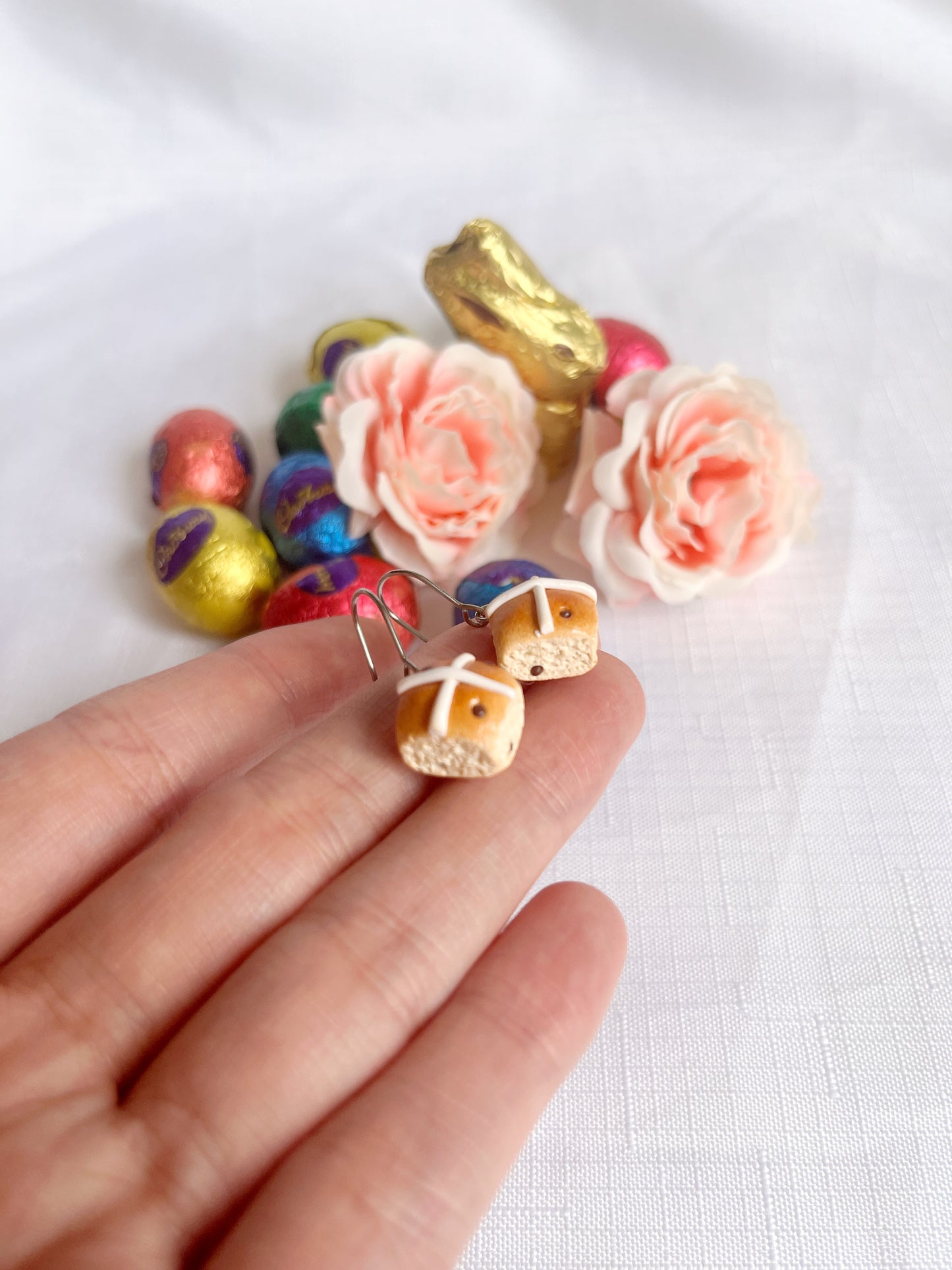 A hand displays two Hot Cross Bun Polymer Clay Earrings by silverstar.nz. Nearby, pastel eggs, pink roses, and a golden bunny adorn a white background. These handmade earrings offer a unique, playful accent to your Easter look.