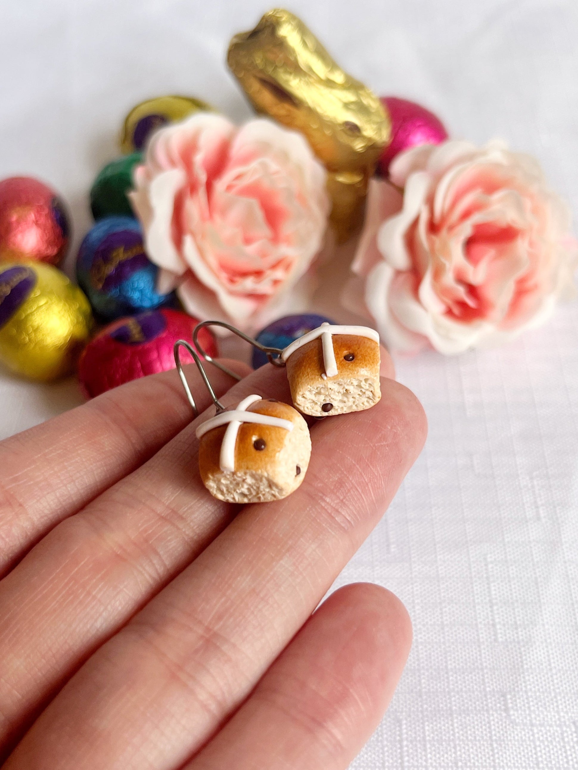 A hand displays unique Hot Cross Bun Polymer Clay Earrings by silverstar.nz. In the background, colorful chocolate eggs, a gold-wrapped chocolate bunny, and pink roses are arranged on a white cloth.