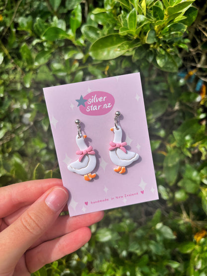 A hand holds a card reading silverstar.nz with Goose with Bow Polymer Clay Earrings, handmade in New Zealand, shaped like white geese with pink bows, set against leafy green foliage.