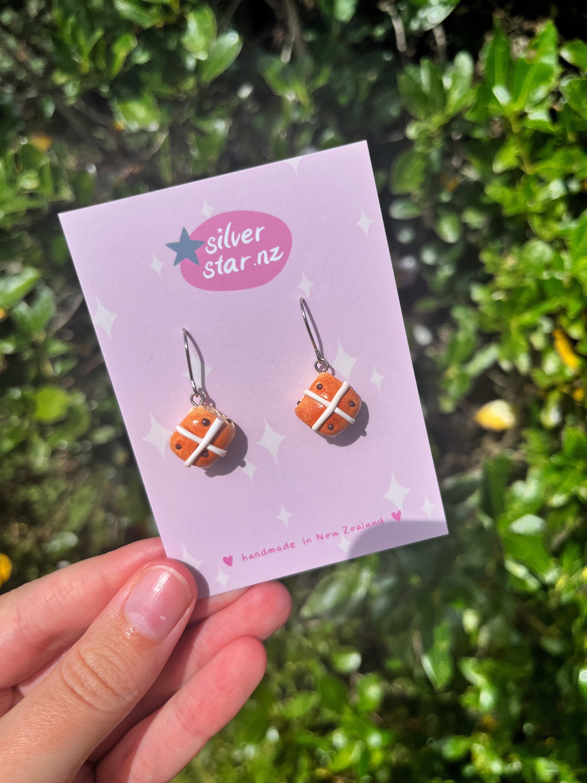 A hand holds a branded silverstar.nz card highlighting Hot Cross Bun Polymer Clay Earrings with stainless steel hooks, handmade in New Zealand. Green foliage is in the background.