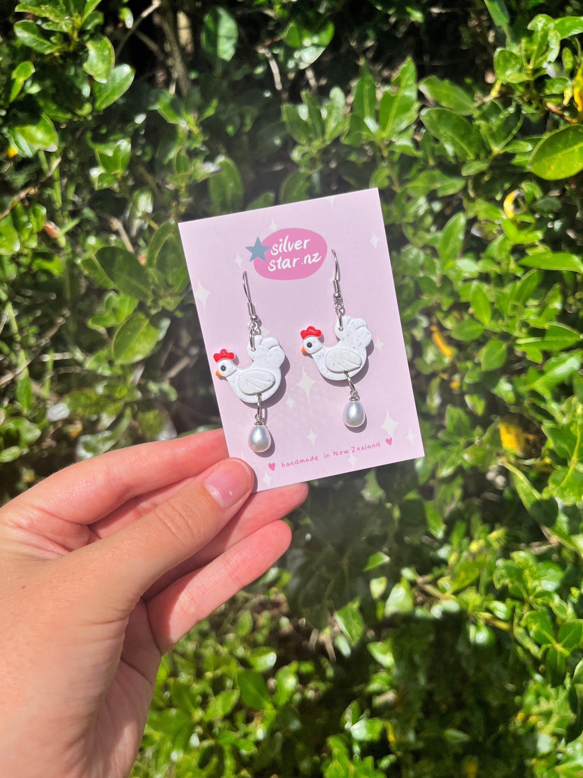 A hand holds a card labeled silverstar.nz displaying Chicken with Egg Polymer Clay Earrings—white chicken shapes with pearl-like drops and stainless steel hooks—handmade in New Zealand, with green foliage in the background.