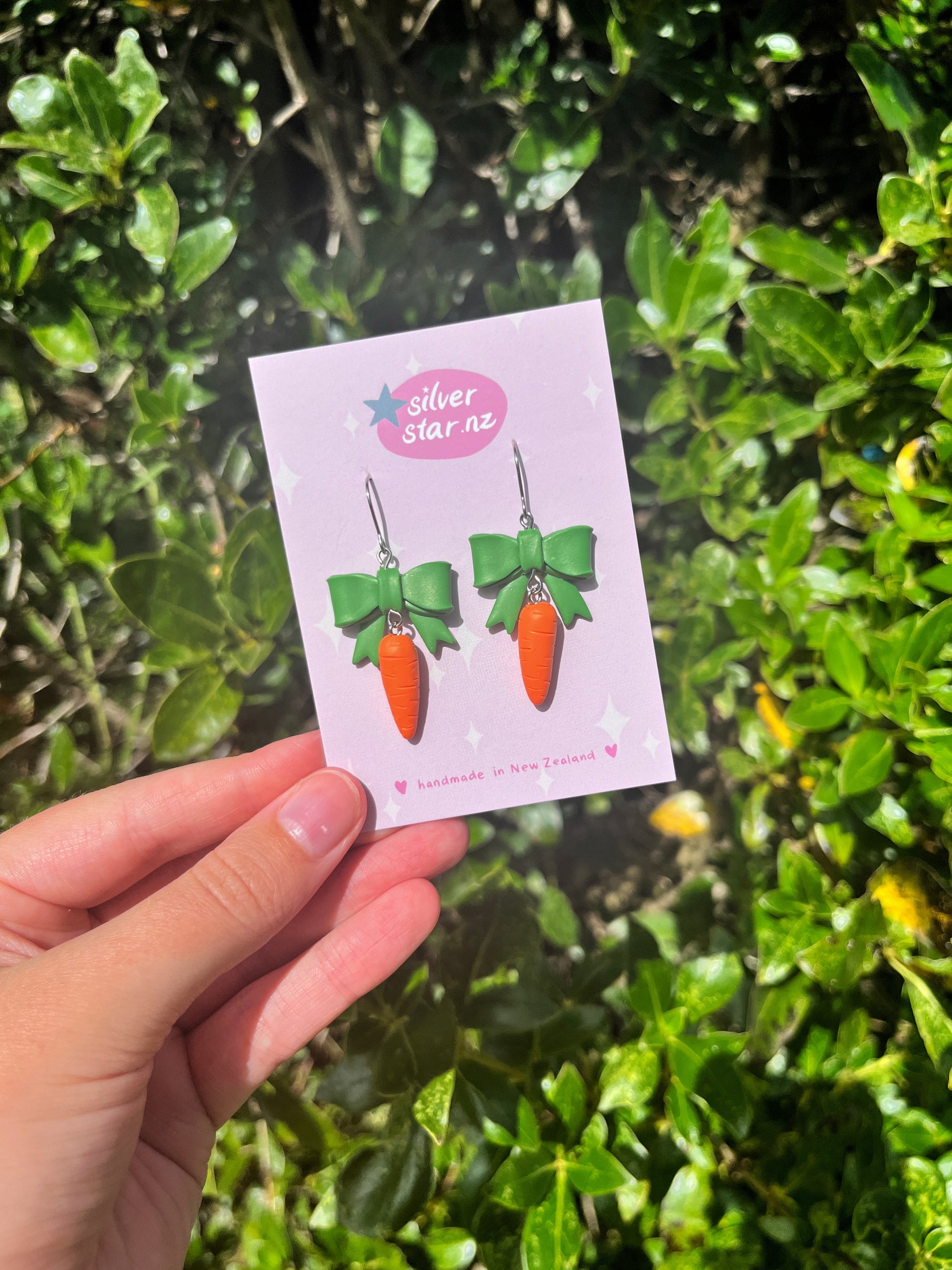 A person holds Carrots with a Bow Polymer Clay Earrings by silverstar.nz, featuring green ribbons and displayed on a pink card against lush green foliage.