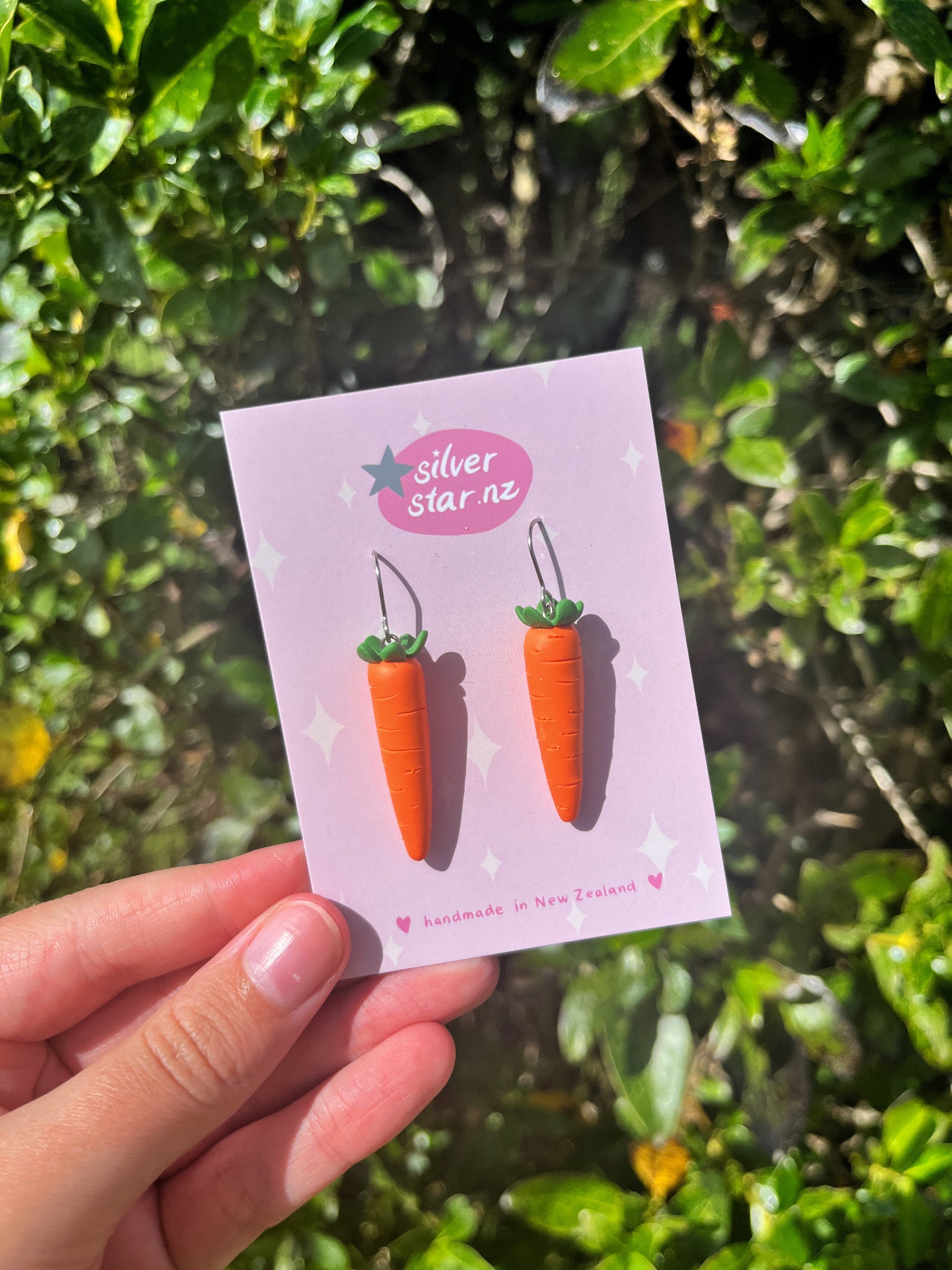 A hand displays the Carrot Polymer Clay Earrings by silverstar.nz on a card with stars and hearts. Featuring green tops and long orange bodies, these handmade earrings are set against leaves, showcasing their New Zealand craftsmanship.