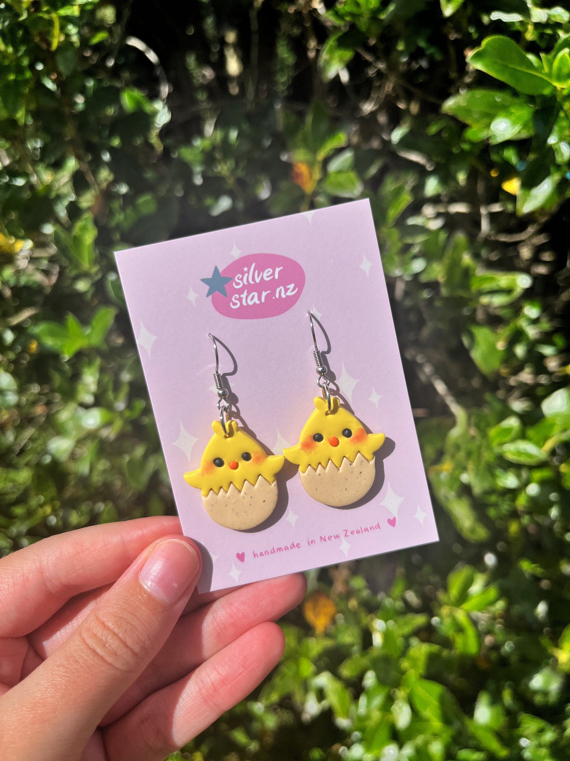 A hand holds a card labeled silverstar.nz with the Egg Baby Chick Polymer Clay Earrings, showing handmade earrings shaped like yellow chicks hatching from eggs, against a backdrop of lush green foliage.
