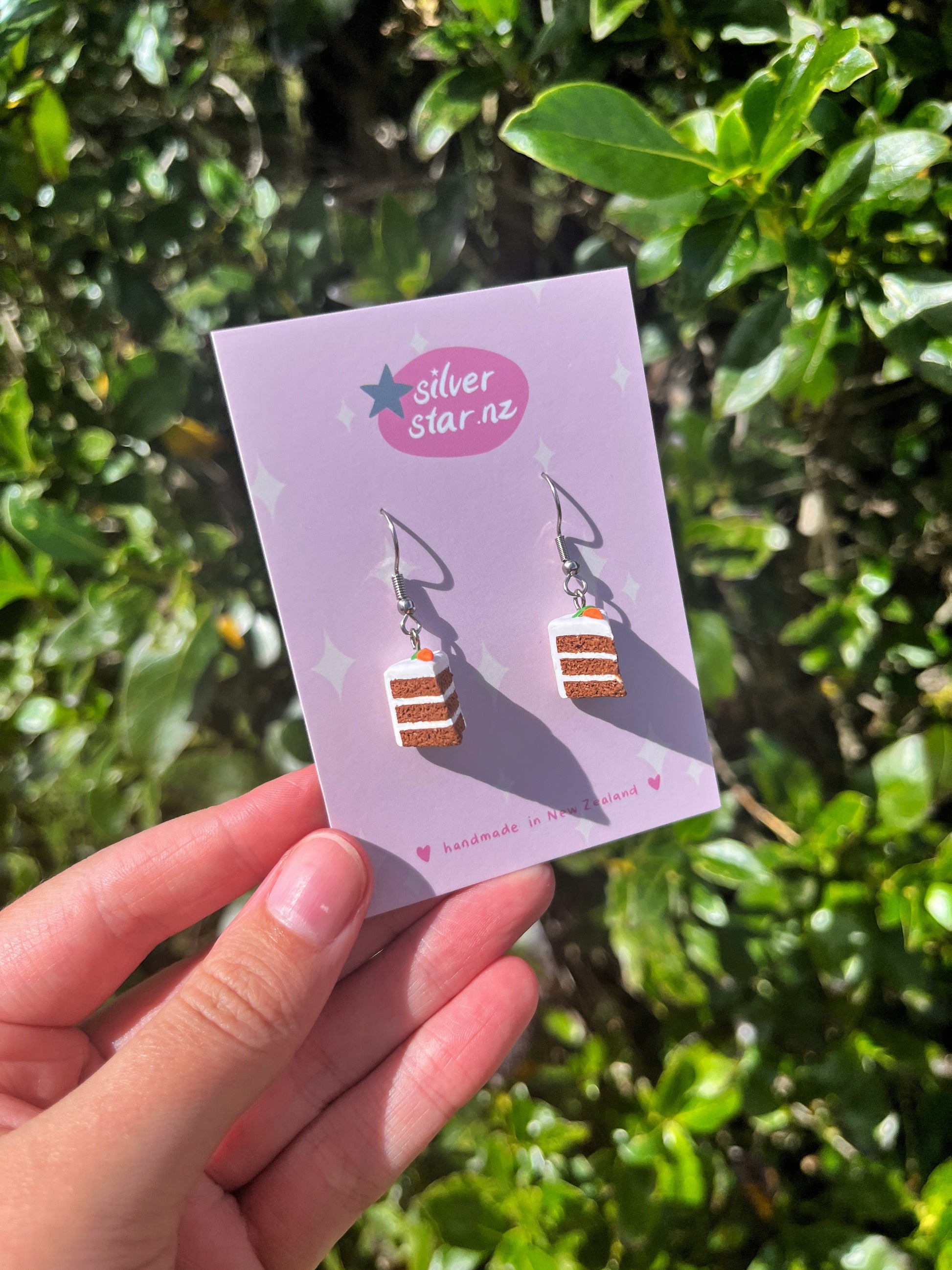 A hand displays the Carrot Cake Polymer Clay Earrings by silverstar.nz, featuring handmade dangle earrings shaped like layered cakes in brown, pink, and white stripes with stainless steel hooks. Vibrant green leafy plants provide a fresh background.