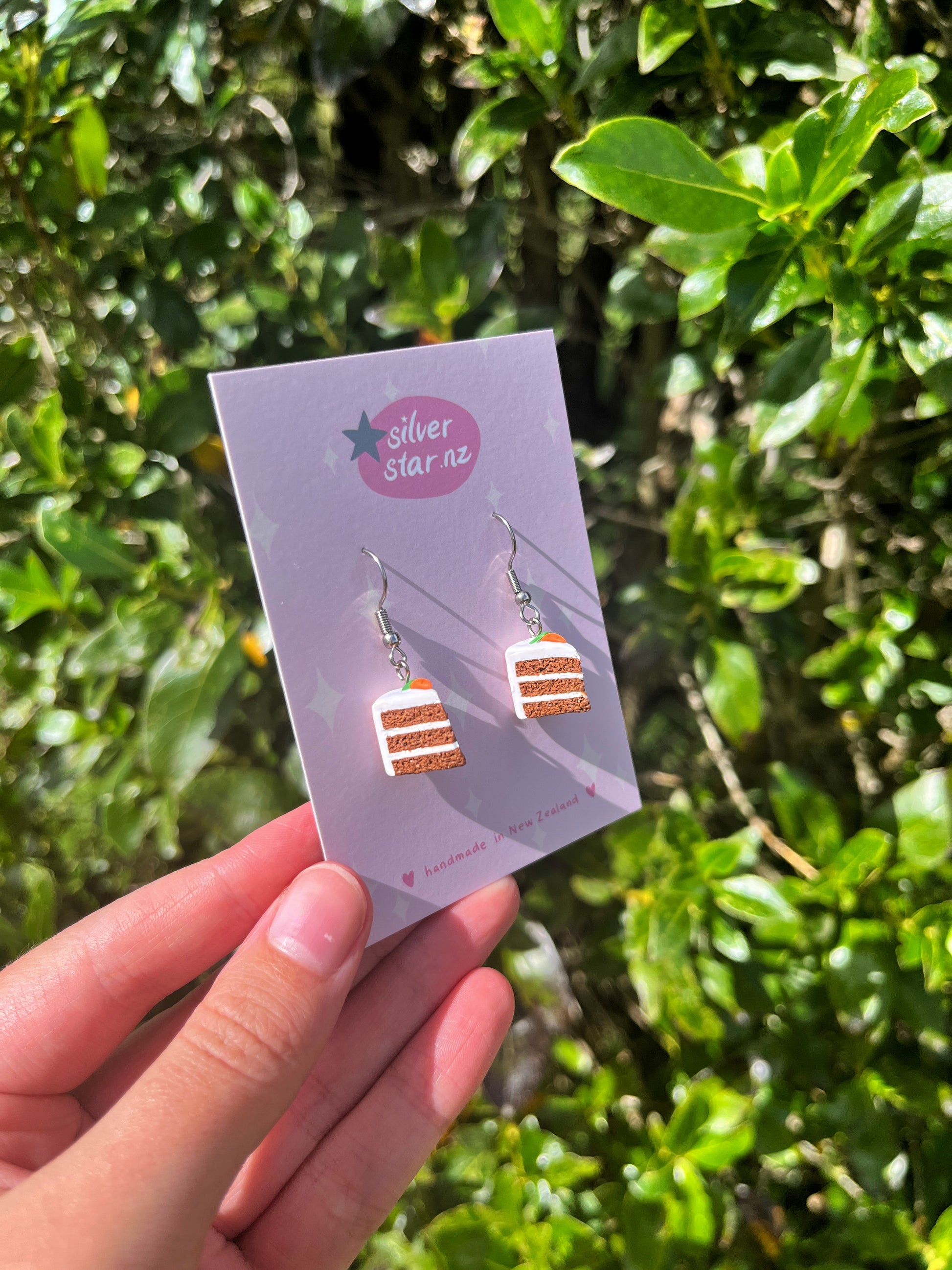 A hand holds a card featuring Carrot Cake Polymer Clay Earrings with pink frosting and orange layers from silverstar.nz. The card reads handmade in New Zealand, with stainless steel hooks, set against a green foliage backdrop.