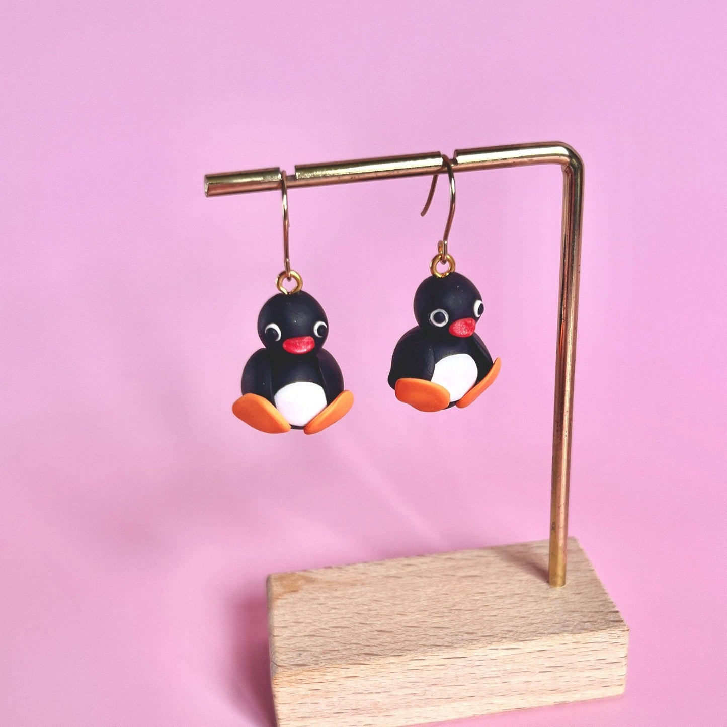 Displayed against a pink background, the Pingu Polymer Clay Earrings dangle elegantly from a gold stand. Each handcrafted earring showcases a detailed black and white penguin with orange beak and feet, suspended from secure stainless steel hooks.