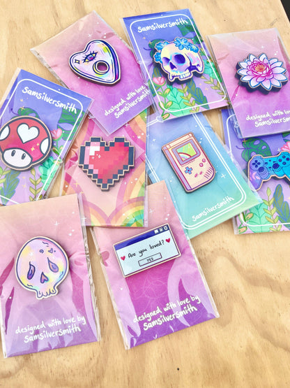 A vibrant assortment of enamel pins is displayed on cherry veneered MDF, featuring artist-designed creations such as a mushroom, heart, skull, flower, game console, ghost, and a pin with the message "Are you loved?" Each piece proudly carries the label "designed with love by SamSilverSmith," including the Pixel Heart Wooden Pin.